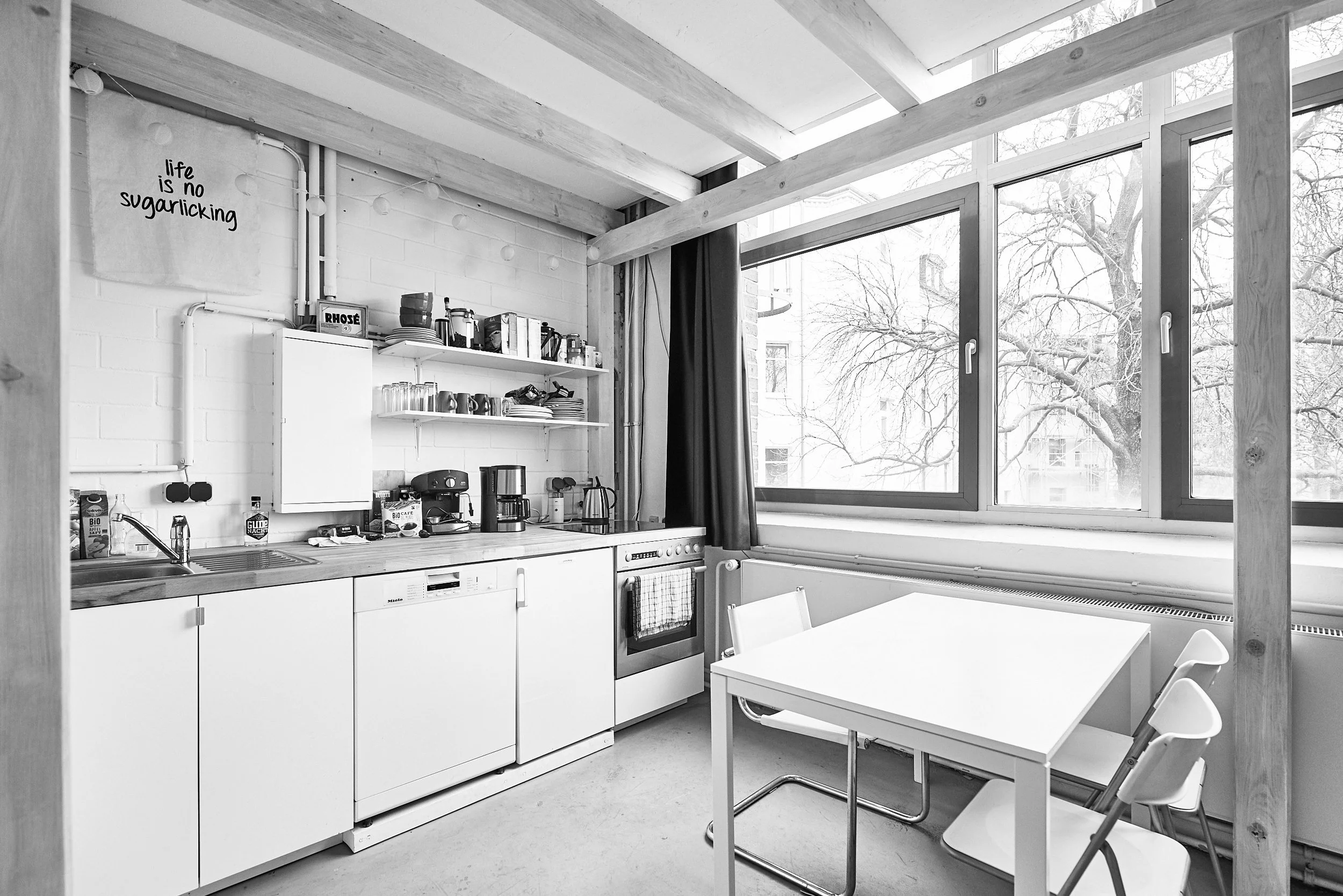 Ein schwarz-weißes Bild einer modernen Küchenzeile mit einem Esstisch und Stühlen neben einem großen Fenster, offene Regale mit Geschirr, Küchenmaschinen und Lebensmitteln, eine weiße Wand mit einem Spruch.