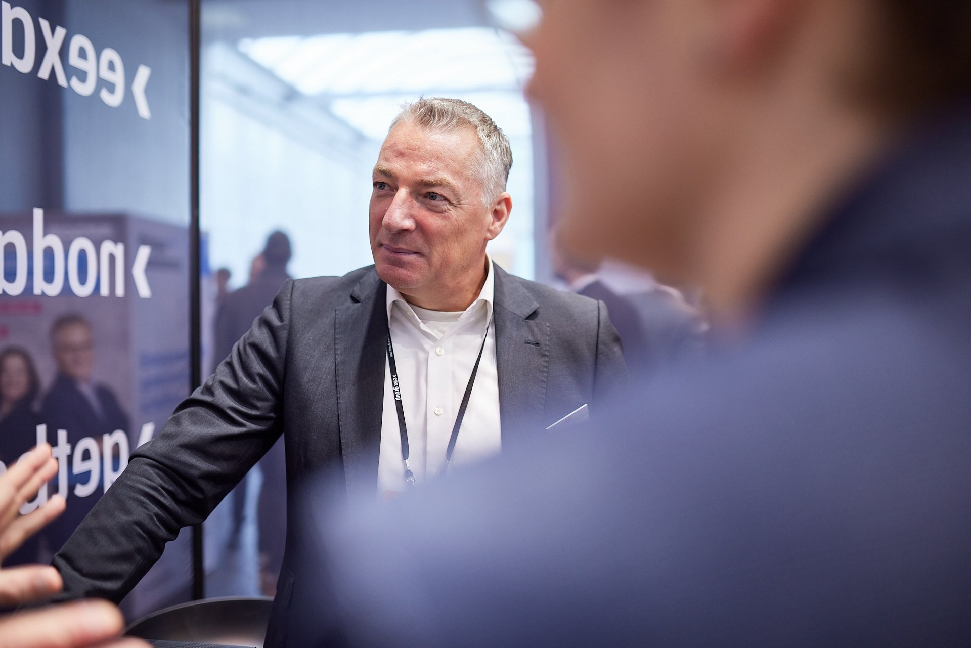 Ein Mann in einem grauen Anzug spricht mit einer Person, nur ein Teil seines Gesichtes ist sichtbar, im Hintergrund sind weitere Menschen und ein Plakat mit der Aufschrift 'Texcon'.