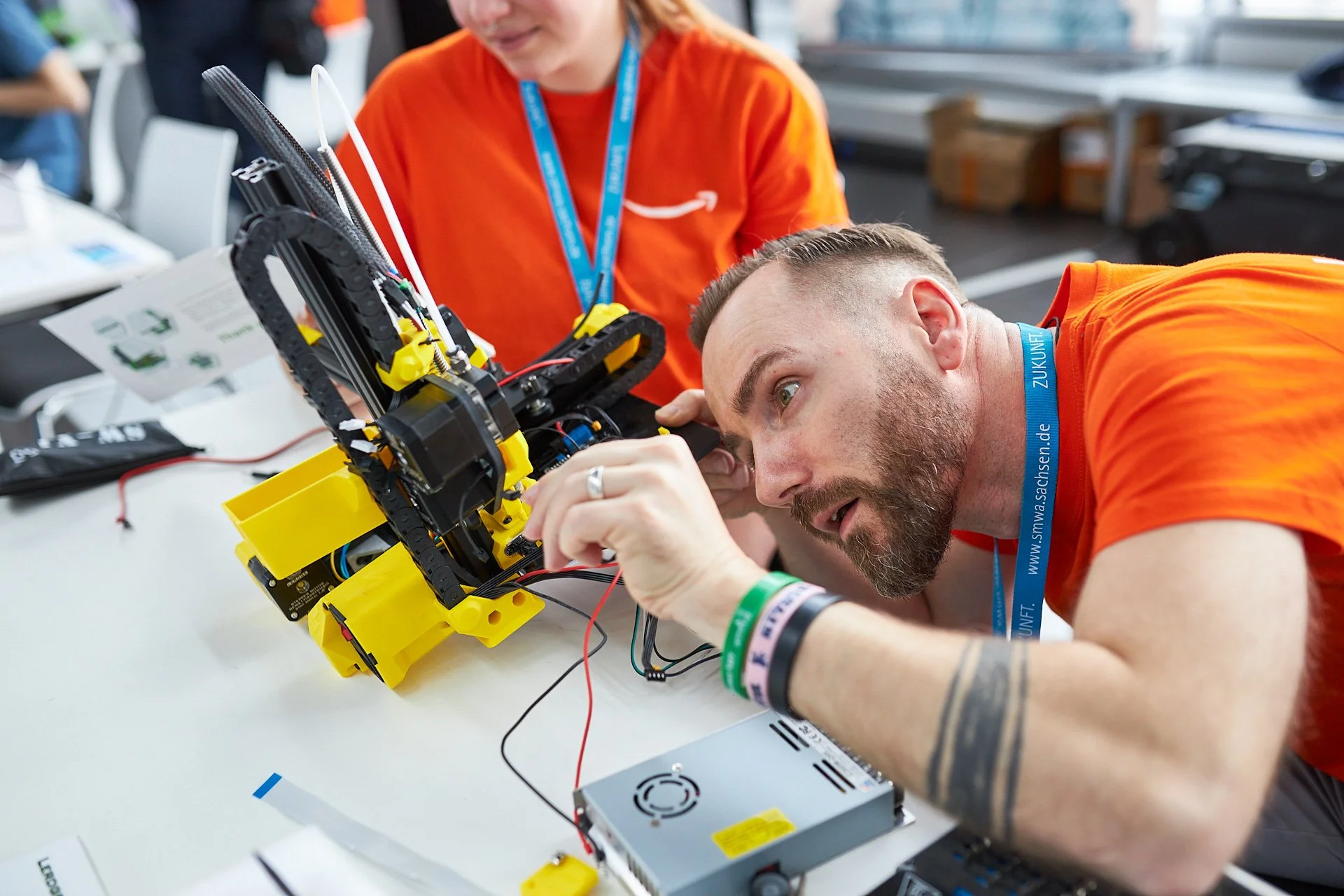 Mann arbeitet an einem Roboter in einem Workshop, trägt ein orangefarbenes T-Shirt und eine blaue Lanyard.
