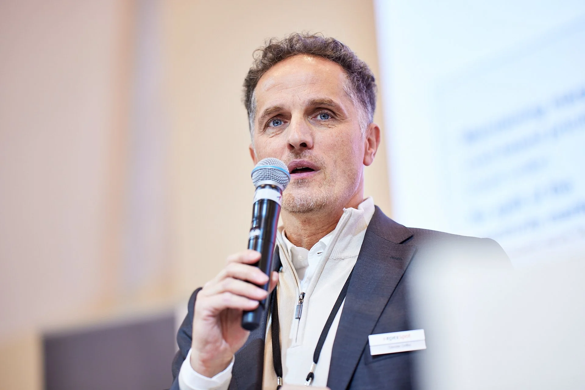 Mann spricht bei einer Konferenz, hält ein Mikrofon in der Hand, trägt einen schwarzen Blazer und eine weiße Zip-Up-Jacke, im Hintergrund eine Präsentationsfolie.