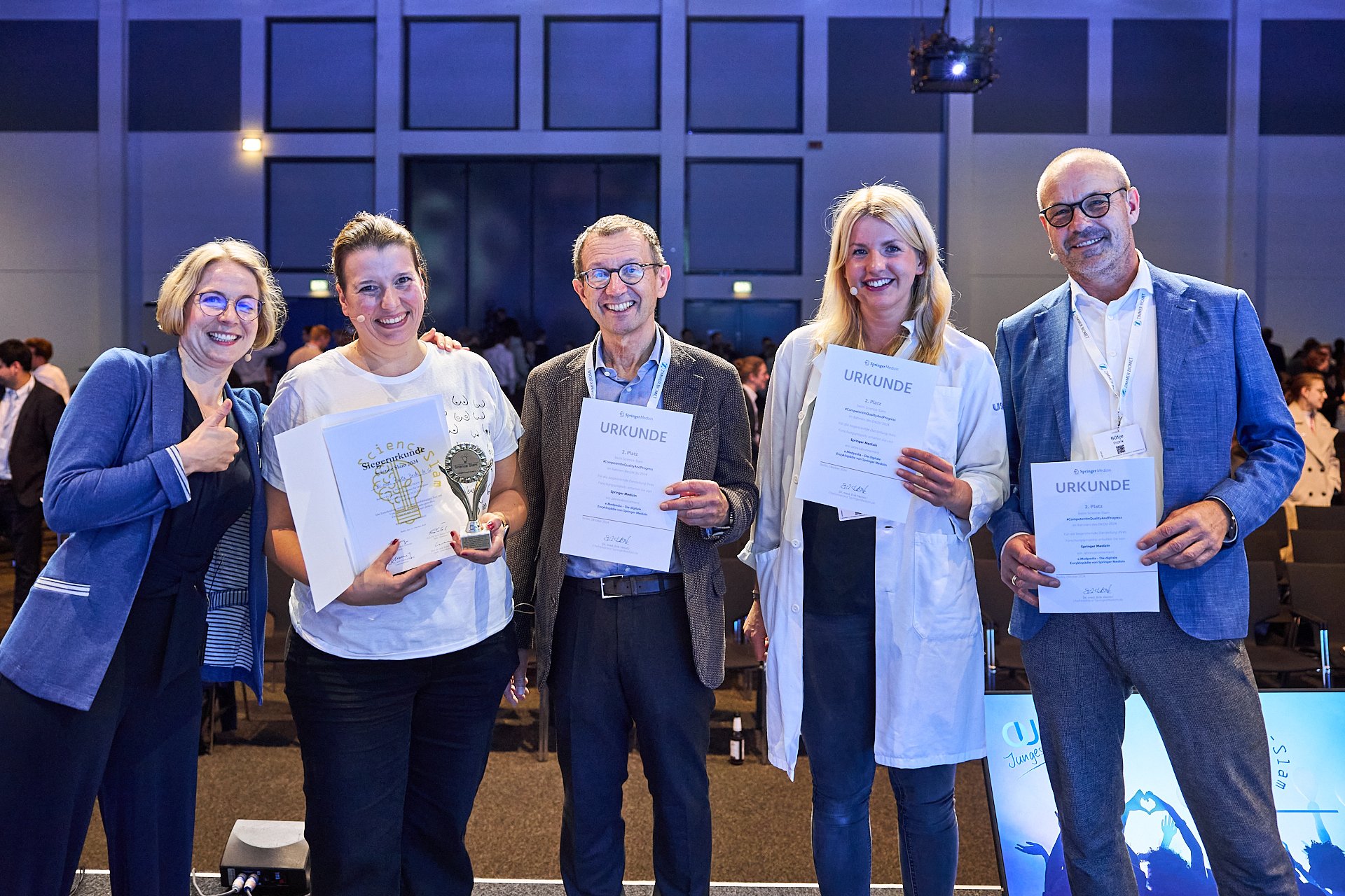 Fünf Menschen auf einer Bühne bei einer Preisverleihung, halten Urkunden und eine Trophäe, alle lächelnd, im Hintergrund weitere Teilnehmer und dunkle Kulisse.