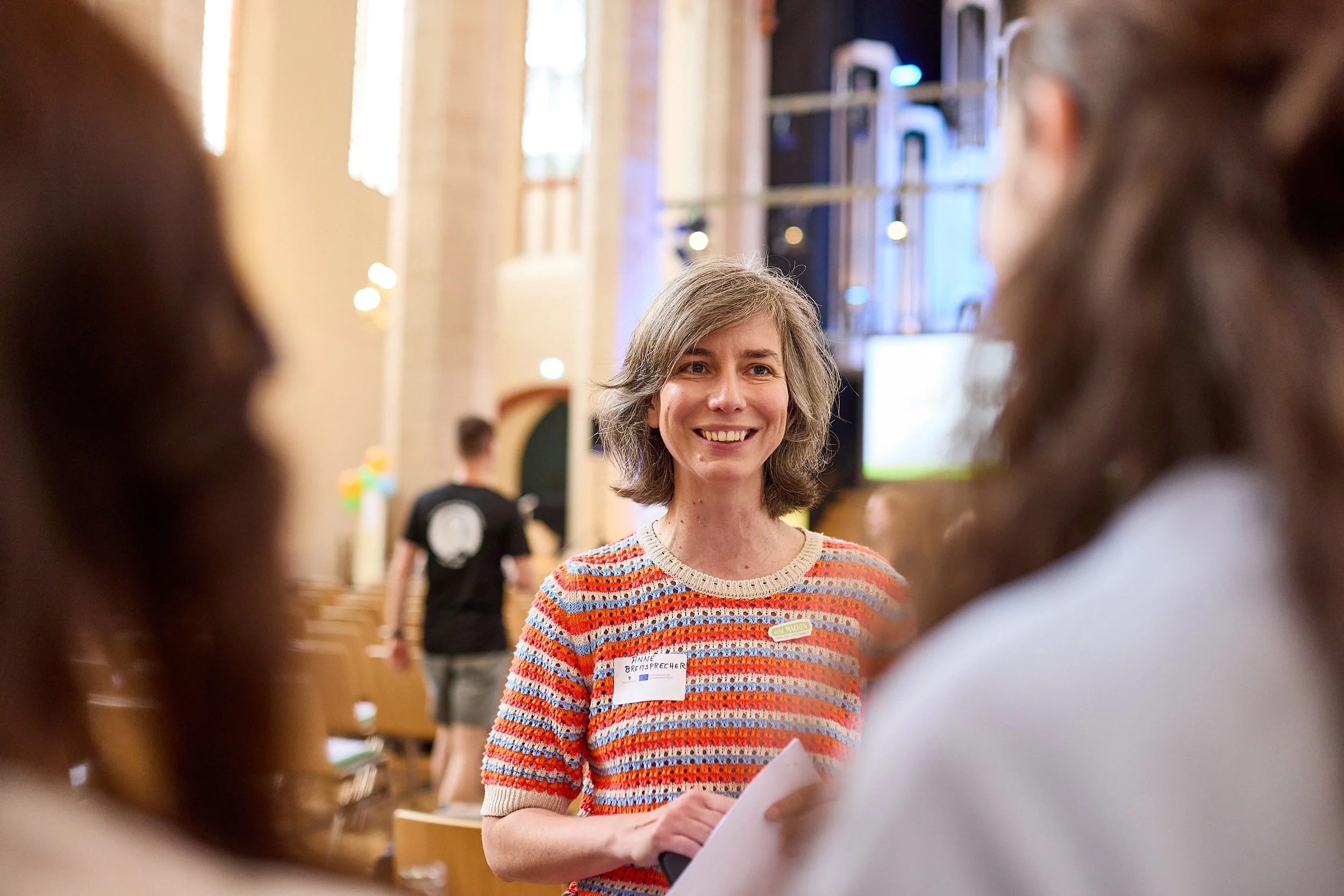 Fröhliche Frau beim Gespräch in einem Raum mit hohen Fenstern und moderner Innenarchitektur, möglicherweise bei einer Veranstaltung oder einem Meeting.