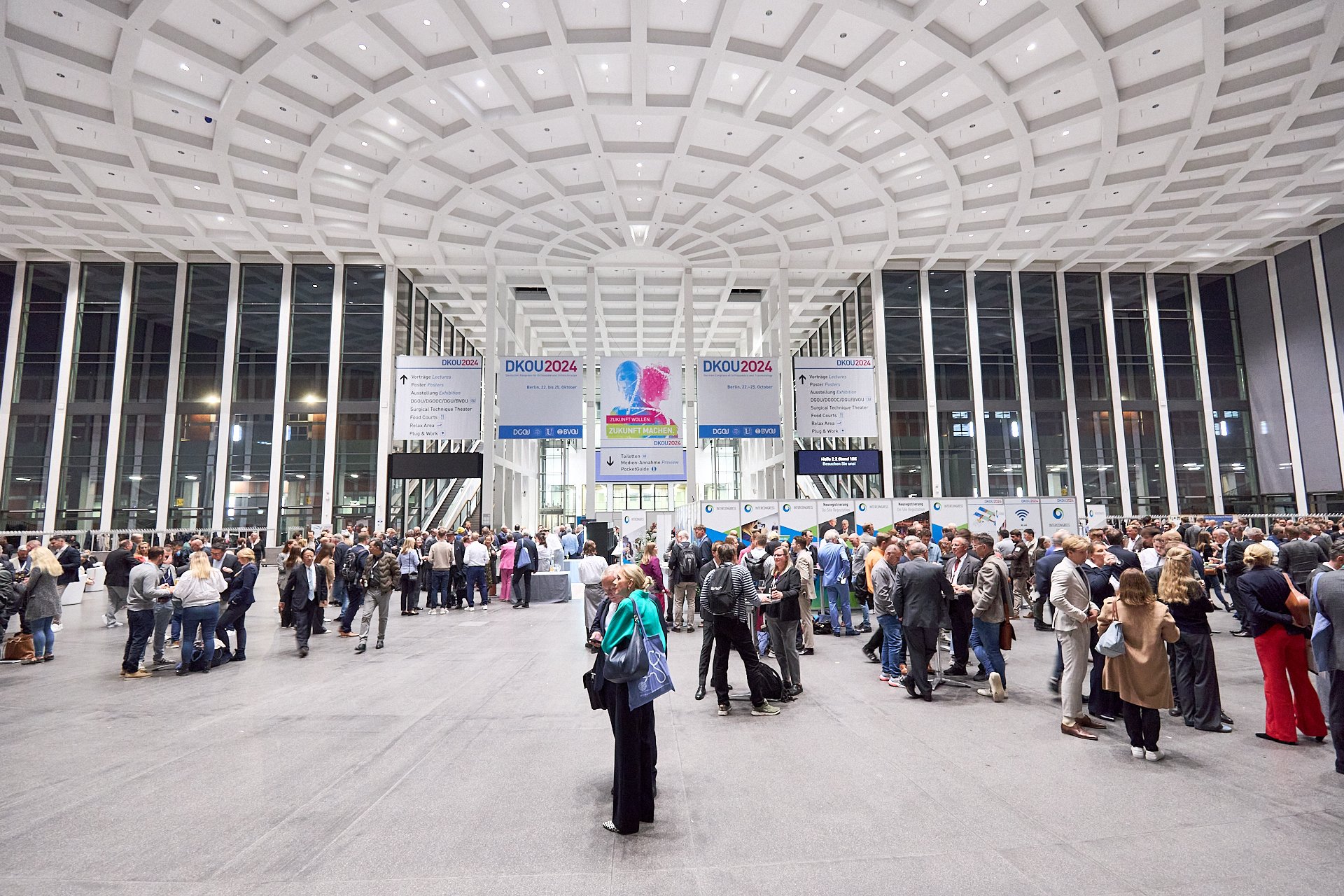 Große Menschenmenge versammelt sich in einer modernen Konferenzhalle mit gerader Deckenstruktur und großen Glasfenstern. Es gibt Informationsstände, digitale Bildschirme und Plakate mit der Aufschrift DKOU 2024.