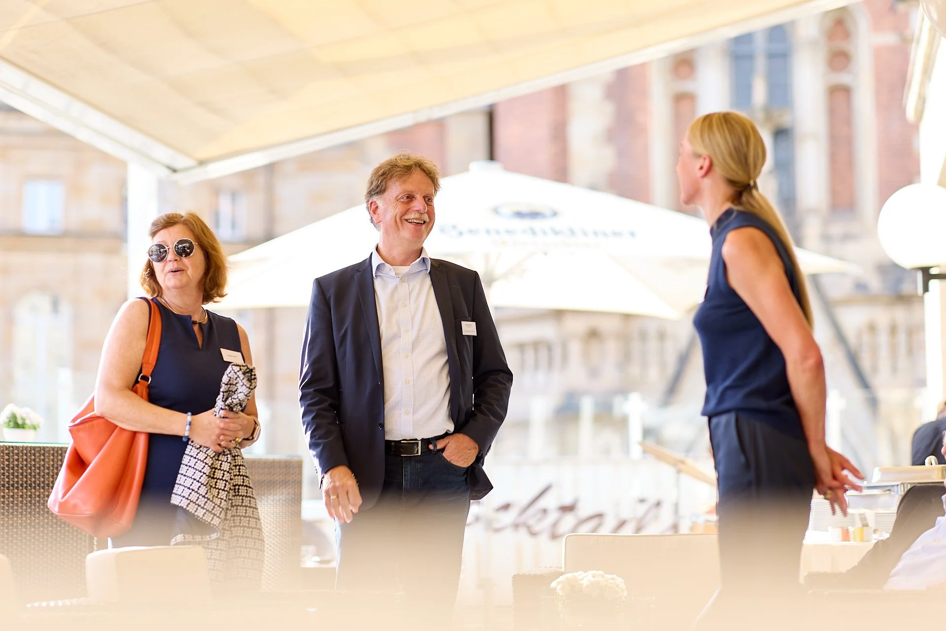 Drei Menschen sprechen sich in einem Restaurant im Freien an. Es sind zwei Frauen und ein Mann. Die Frauen tragen Sonnenbrillen und sind casual gekleidet, der Mann trägt einen Anzug. Im Hintergrund sind Gebäude und Sonnenschirme sichtbar.