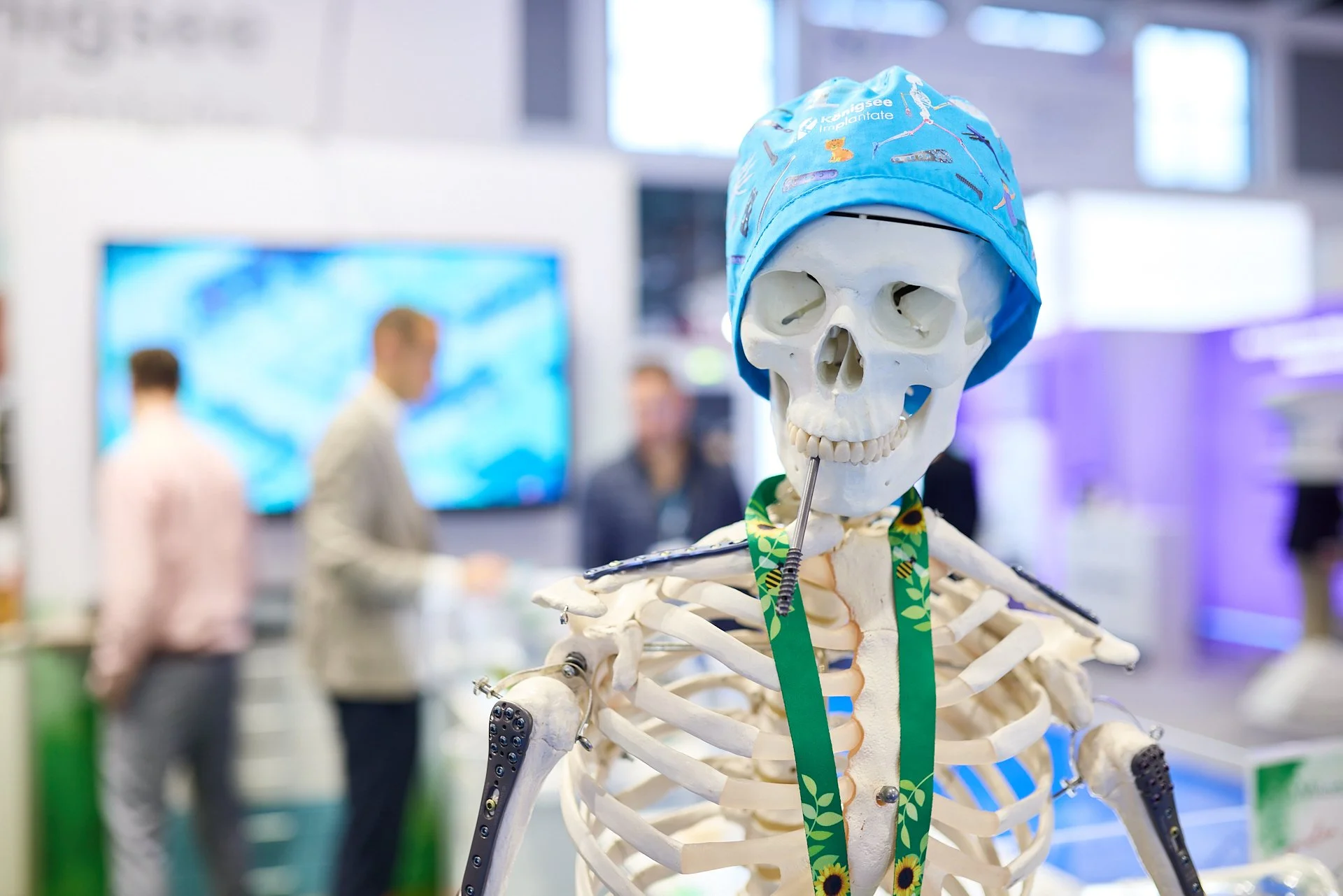 Skelettmodell mit blauer Operationsmütze und Löffel im Mund, auf einer Messe oder Konferenz mit Menschen im Hintergrund.