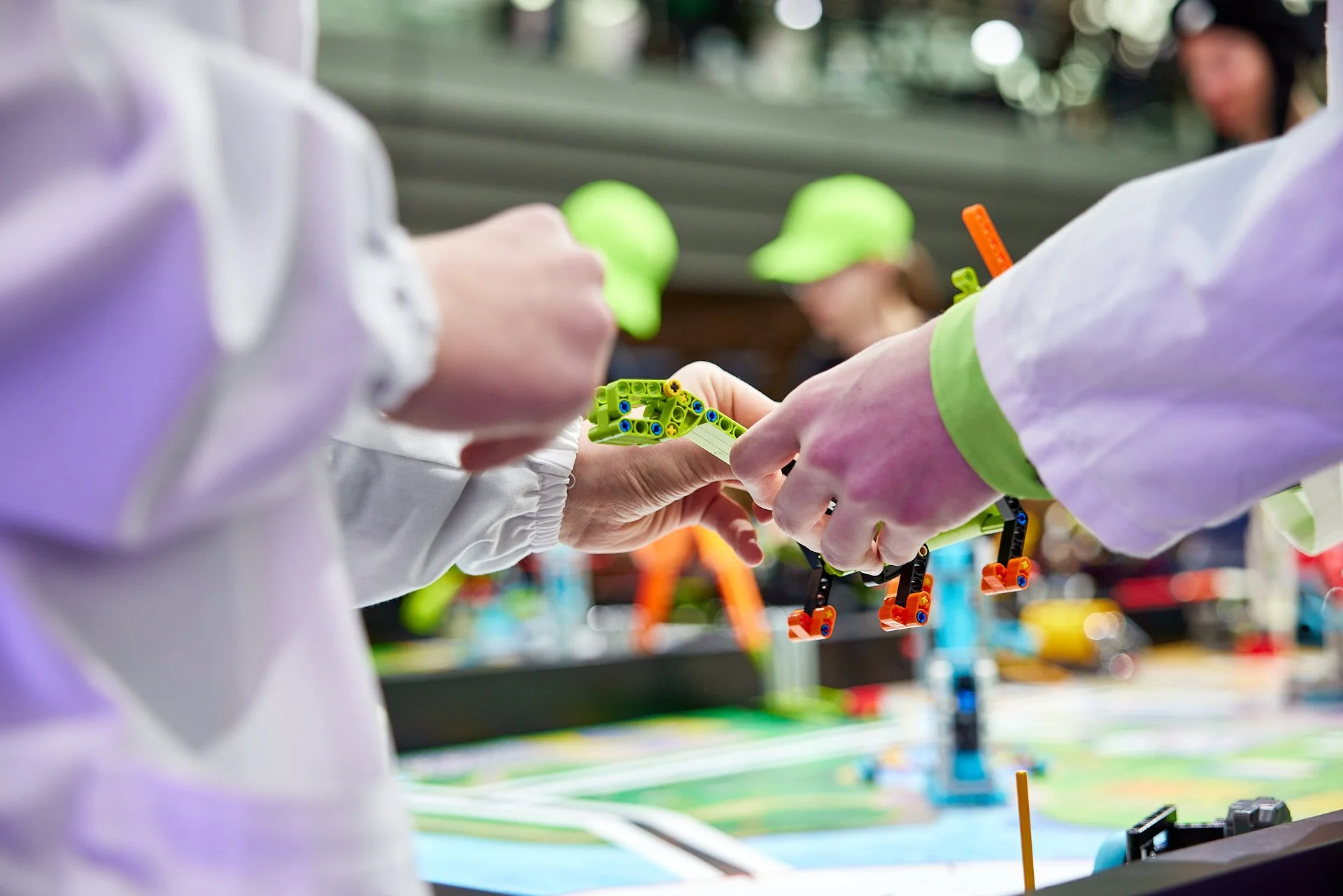 Zwei Personen tauschen eine kleine grüne LEGO-Kreation aus, während sie mit anderen LEGO-Bausteinen und Spielmaterialien auf einem Spieltisch spielen.