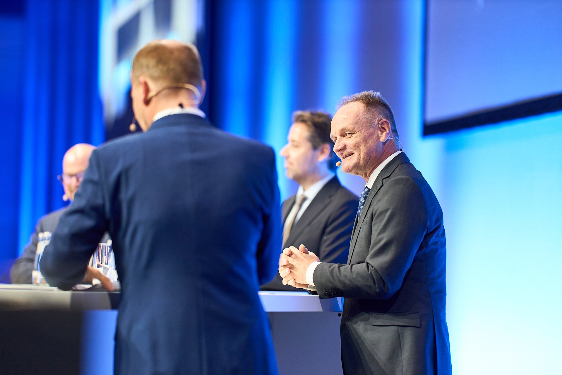 Drei Männer in dunklen Anzügen stehen an einem Podium bei einer Konferenz. Der Mann auf der rechten Seite lächelt und schaut nach oben, während die anderen beiden Männer mit den Rücken zum Bild sprechen.