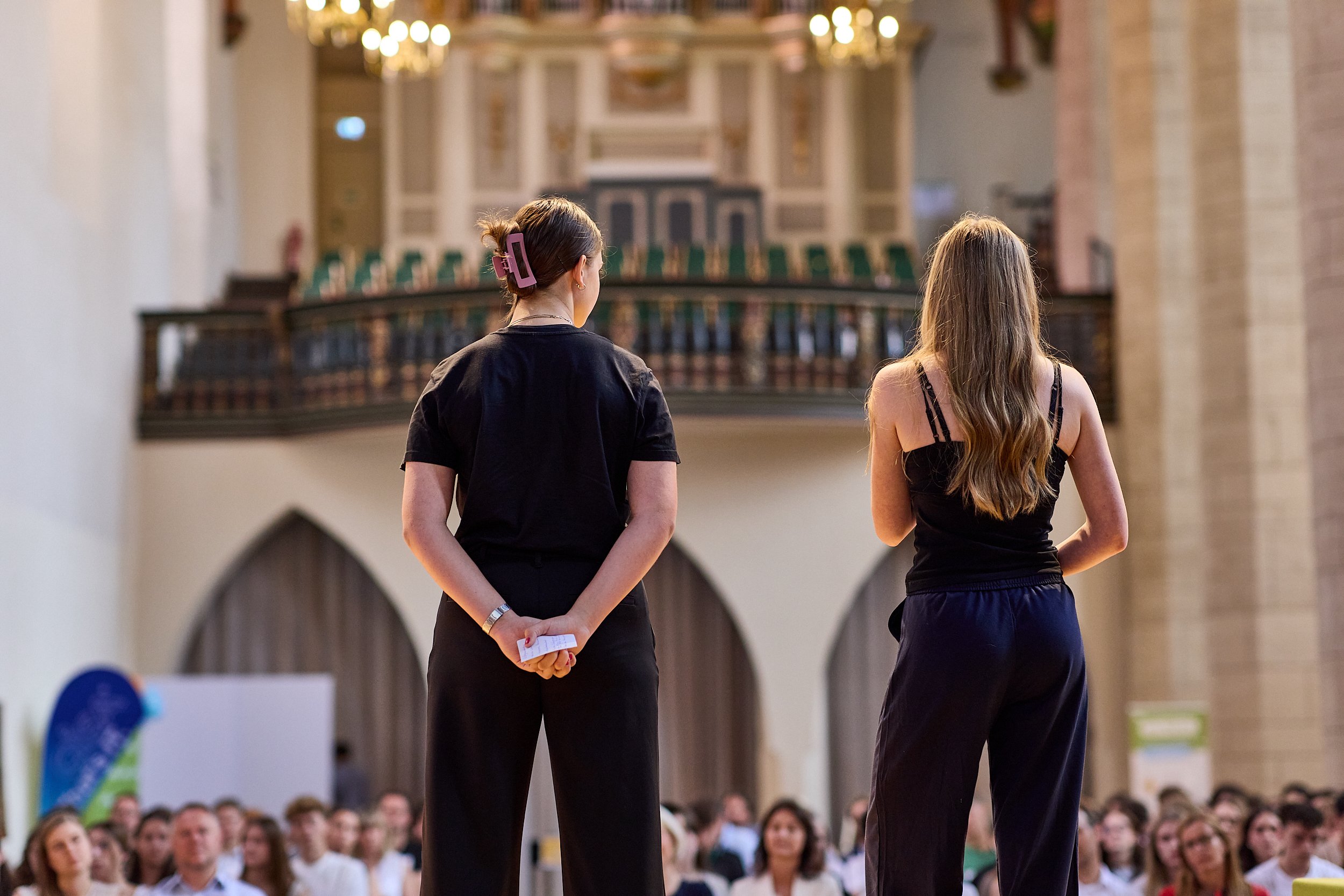 Zwei Frauen stehen auf einer Bühne vor einem Publikum in einem großen Raum, vermutlich einer Kirche oder einem historischen Gebäude.
