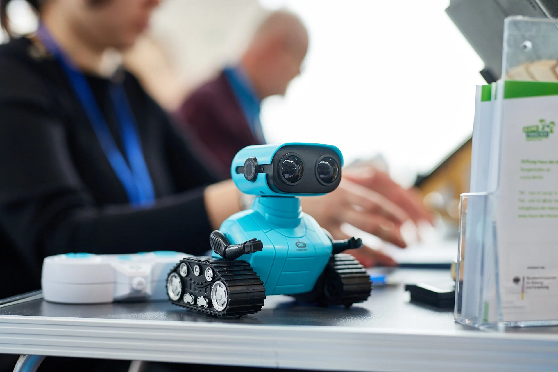 Ein kleiner blauer Roboter mit Rädern sitzt auf einem Tisch, im Hintergrund sind zwei Menschen, die an Laptops arbeiten.