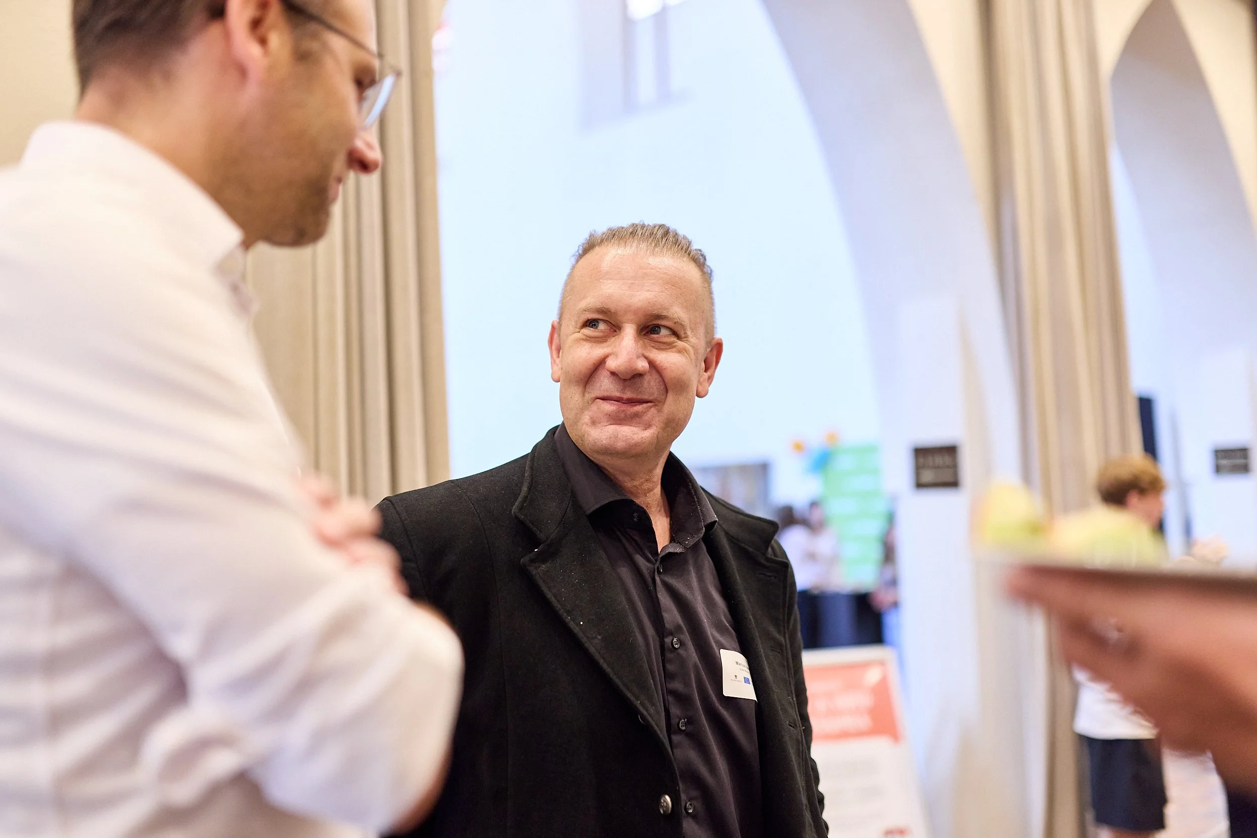 Zwei Männer im Gespräch in einem hellen, offenen Raum mit hohen Bögen, im Hintergrund weitere Personen und Schilder.