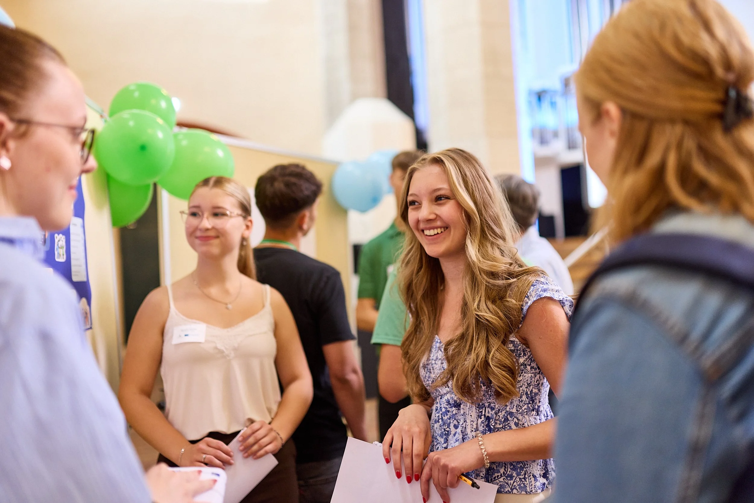 Mehrere junge Menschen im Gespräch auf einer Veranstaltung, mit bunten Ballons im Hintergrund.