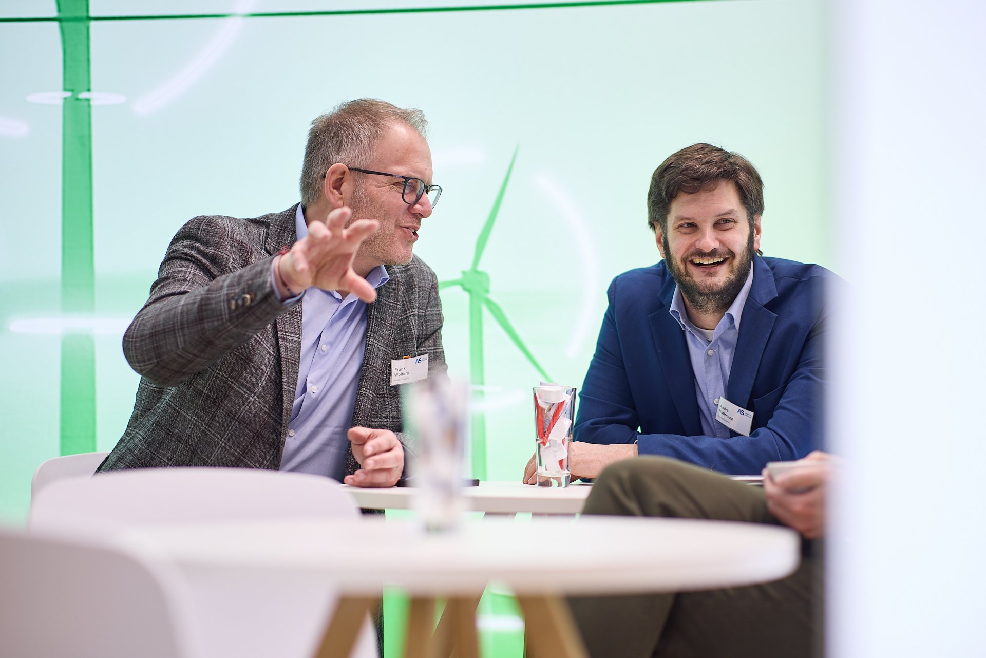 Zwei Männer beim Gespräch, lachen und scherzen bei einer Diskussion in einem modernen Raum, mit einem grünen Windrad im Hintergrund.