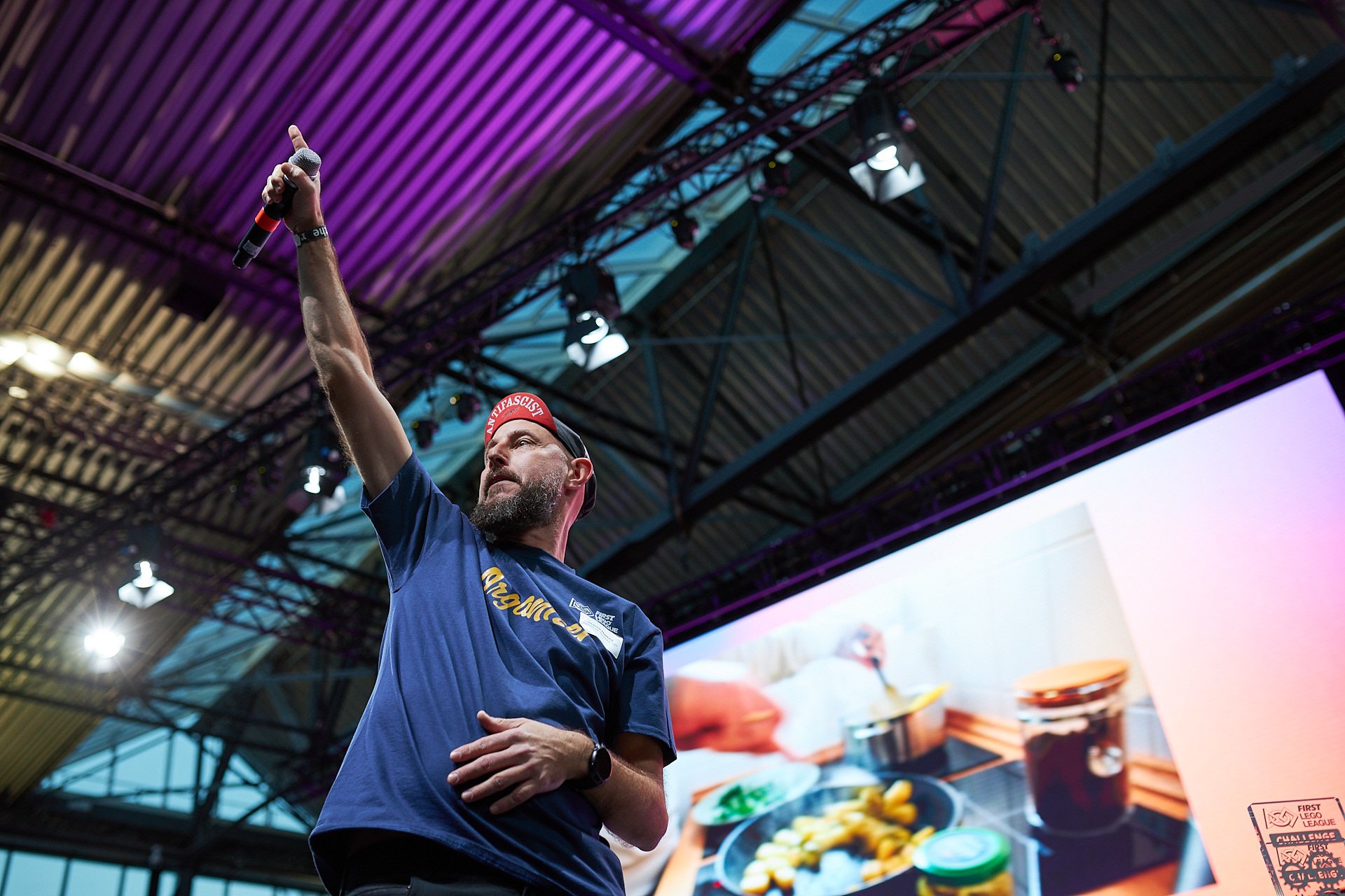 Ein Mann hebt mit einem Mikrofon in der Hand seine rechte Hand in die Luft bei einer Veranstaltung, im Hintergrund eine große Leinwand mit einem Bild von Essen