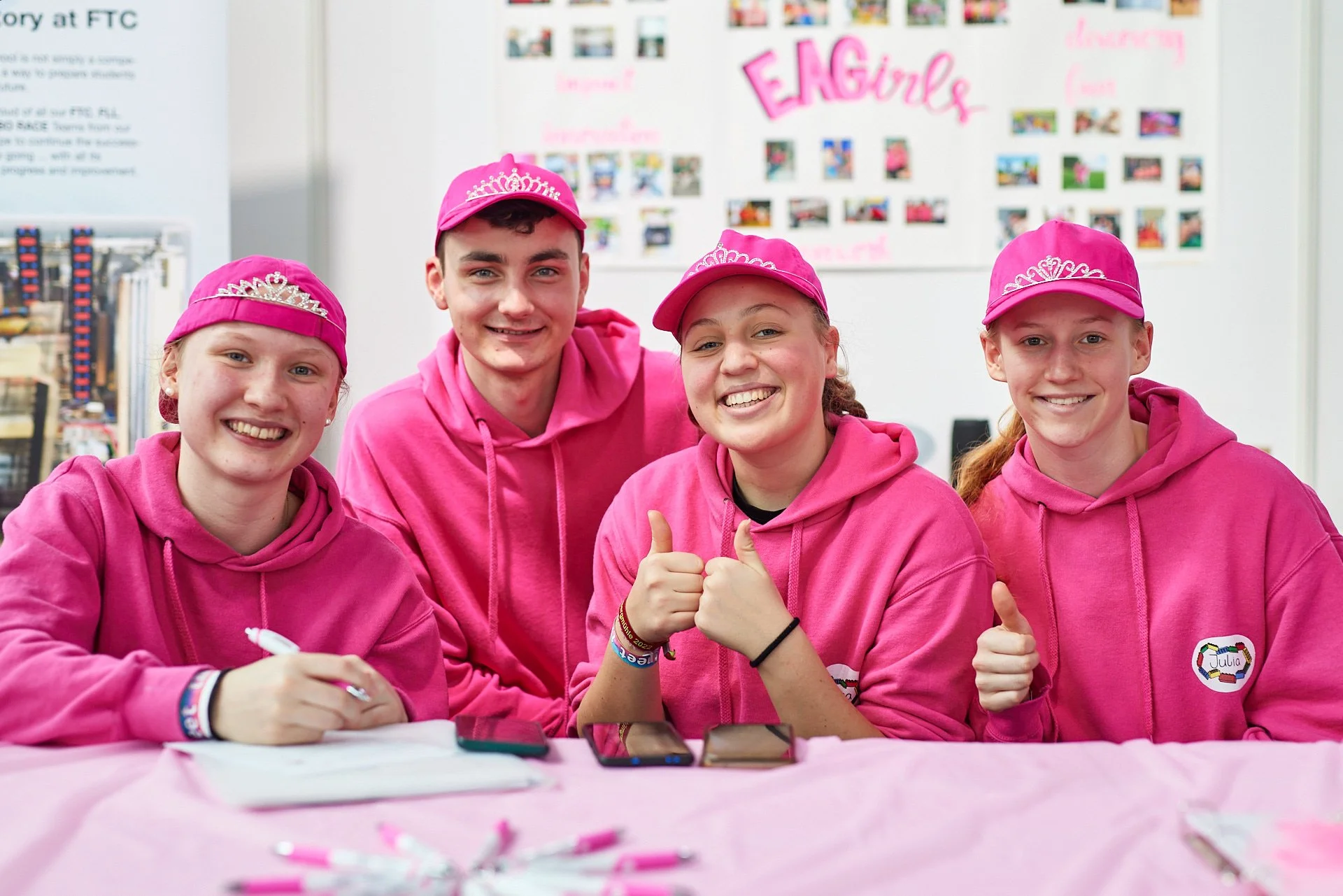 Vier Mädchen in pinken Hoodies und pinken Mützen mit Krönchen sitzen an einem Tisch, lachen und posieren für die Kamera.