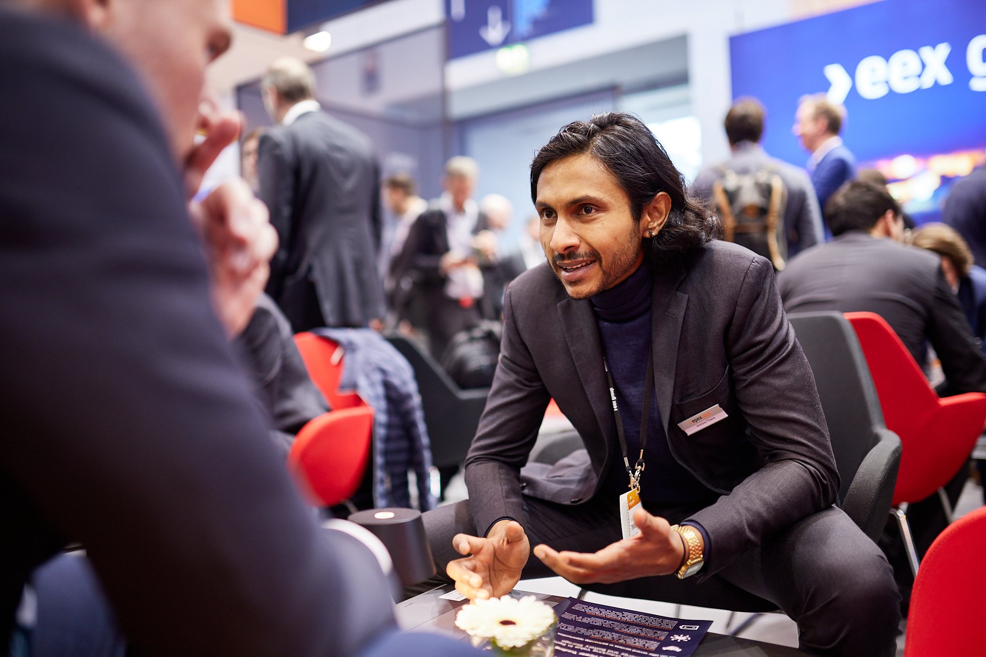 Ein Mann im schwarzen Anzug führt ein Gespräch bei einer Konferenz, im Hintergrund sind weitere Personen und ein großes Display zu sehen.