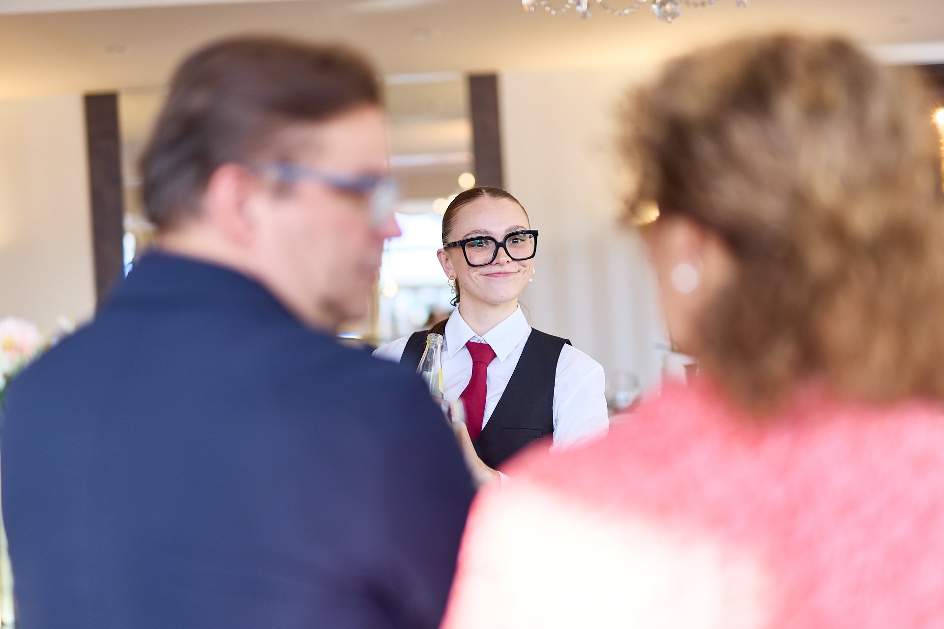 Eine junge Frau mit Brille trägt eine schwarze Weste, weißen Hemd und rote Krawatte und hält ein Getränk in der Hand, während sie mit einem Mann und einer Frau im Gespräch ist.