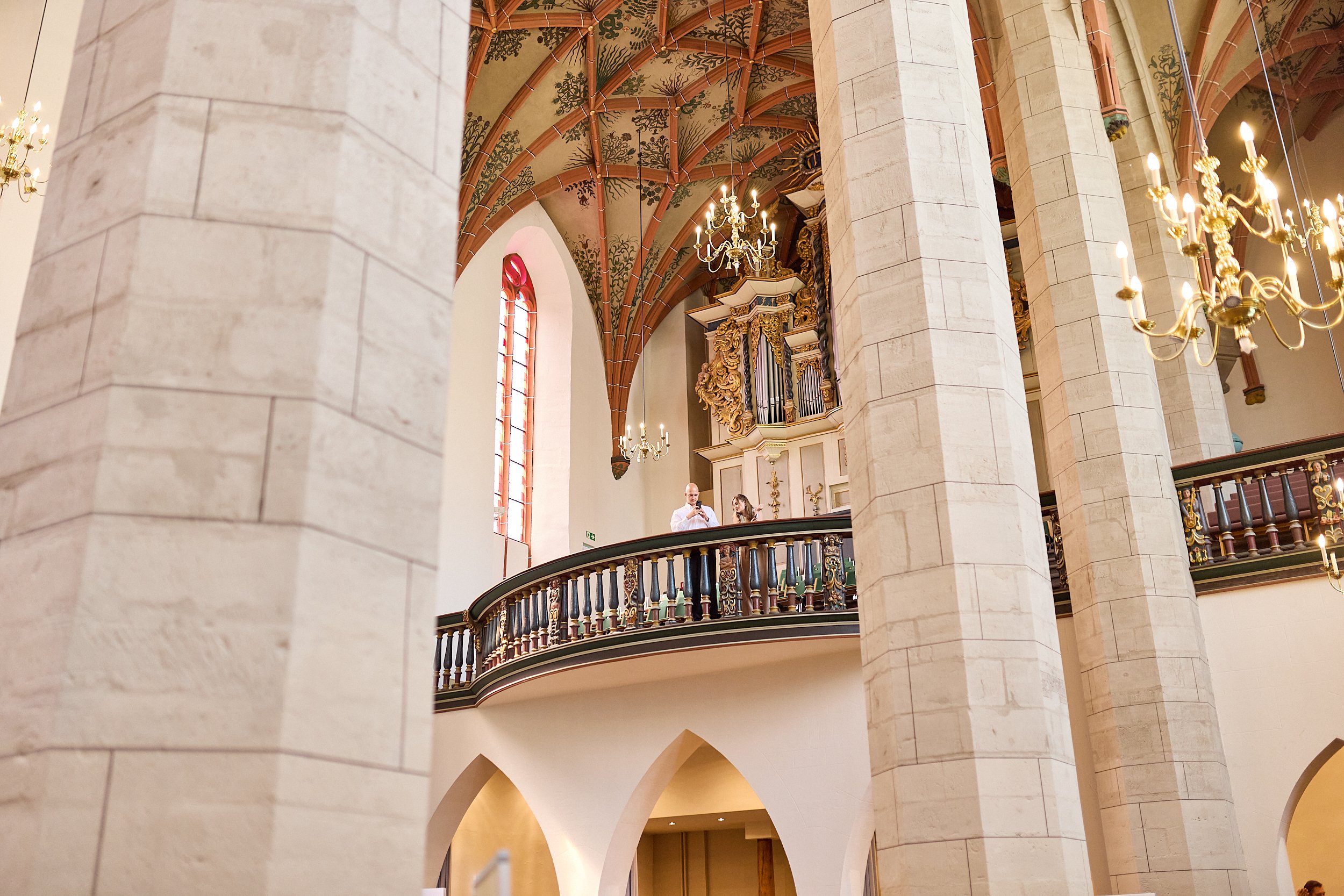 Blick auf die obere Galerie eines gotischen Kircheninneren mit großen Säulen, barocken Kronleuchtern, einem spätbarocken Orgelgehäuse und bunten glasierten Fenster