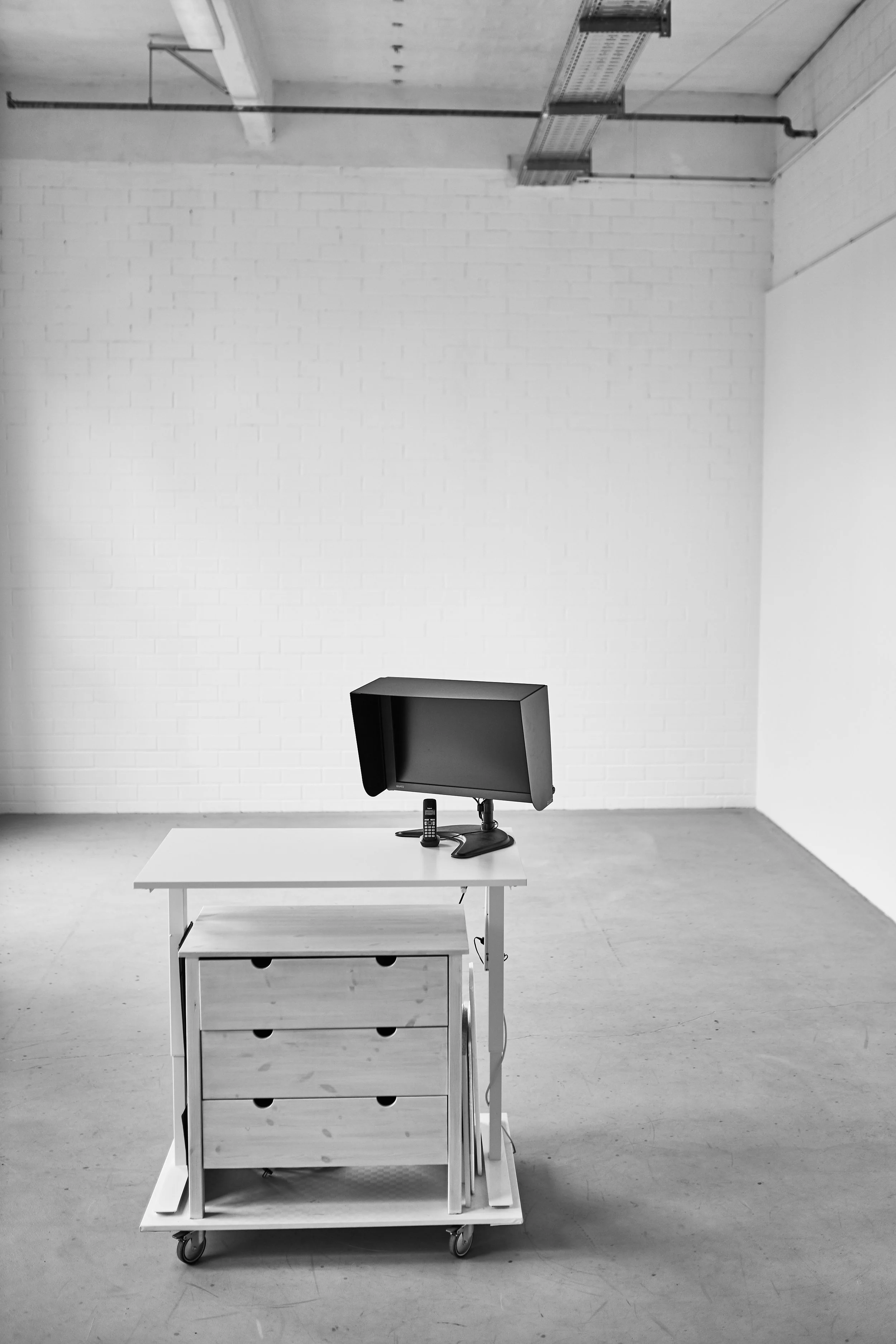 Ein weißer Tisch mit einem Computerbildschirm, einem Telefon und einem Schubladenschrank auf einem Rollwagen in einem leeren Raum mit weißen Wänden und einem Deckenstrahler.