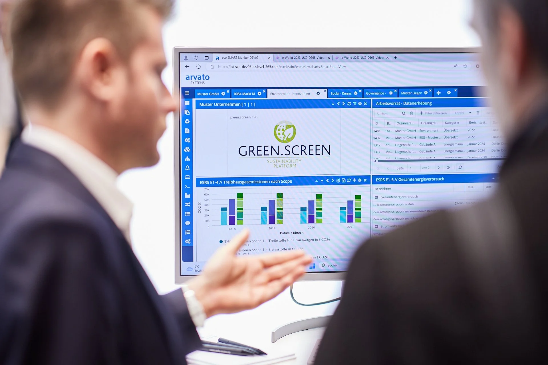 Zwei Personen in einem Gespräch vor einem Bildschirm mit digitalen Diagrammen und Daten, die die Nachhaltigkeitsplattform Green.Screen zeigen.