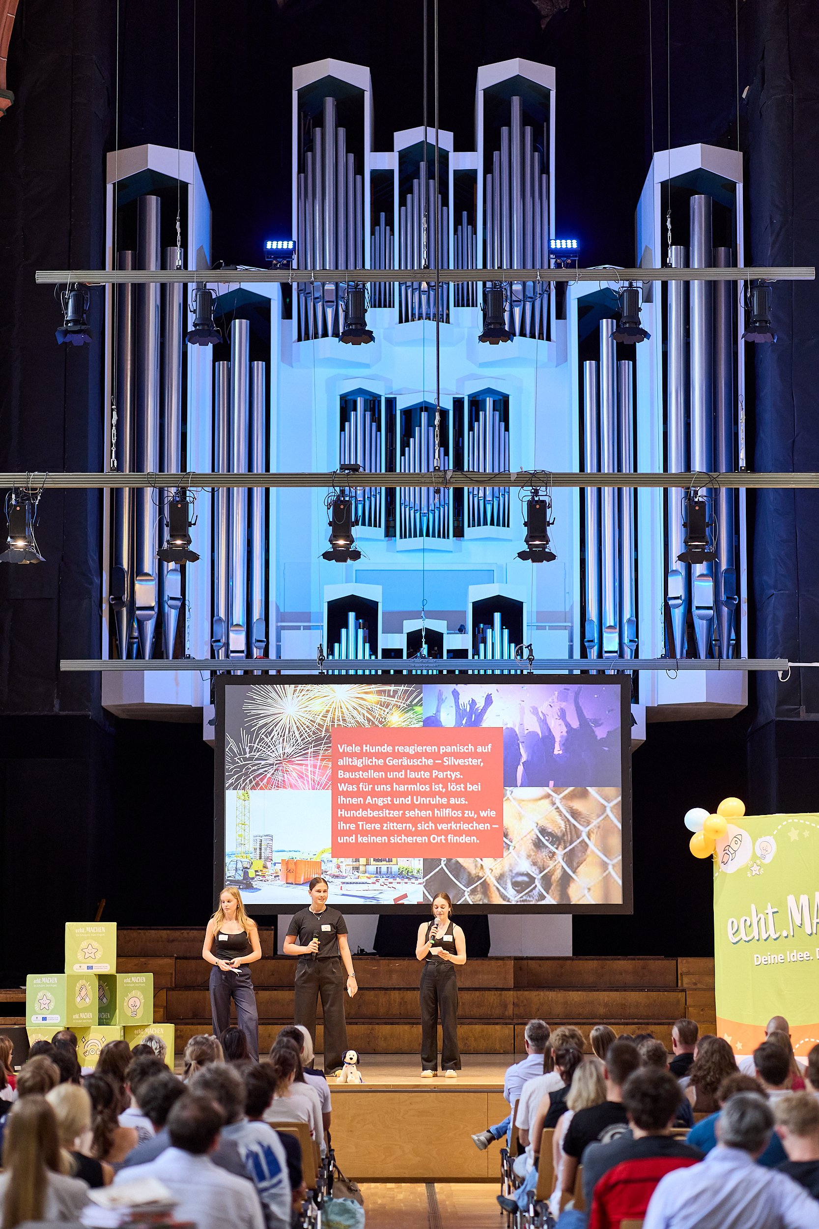 Bühne mit drei Frauen, die vor Publikum sprechen, und großen Bildschirm mit Text und Bildern, im Hintergrund eine Orgel mit vielen Zylindern