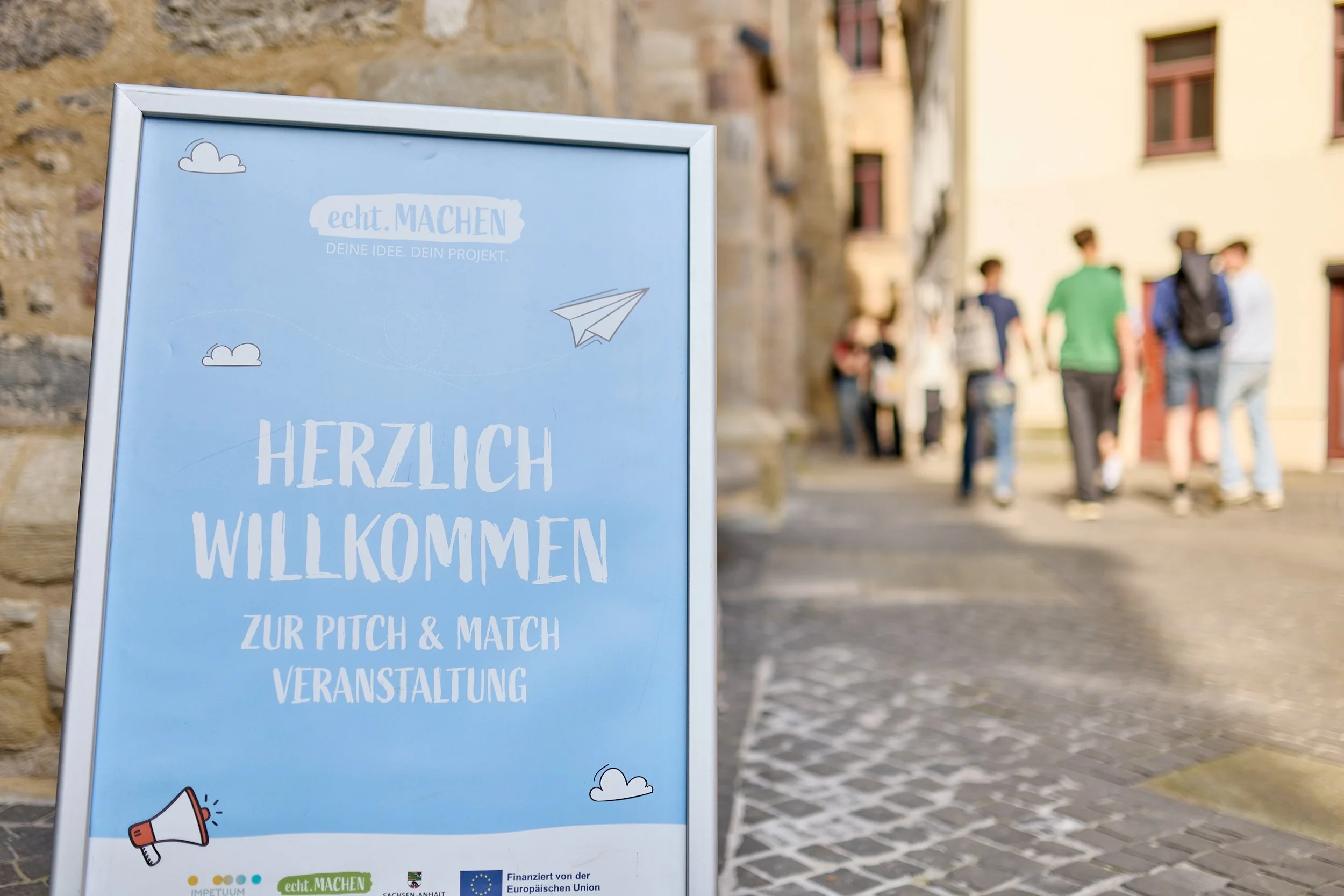 Ein Willkommensschild für eine Pitch & Match Veranstaltung auf einem gepflasterten Platz. Im Hintergrund sind mehrere Personen, die sich unterhalten, sichtbar.