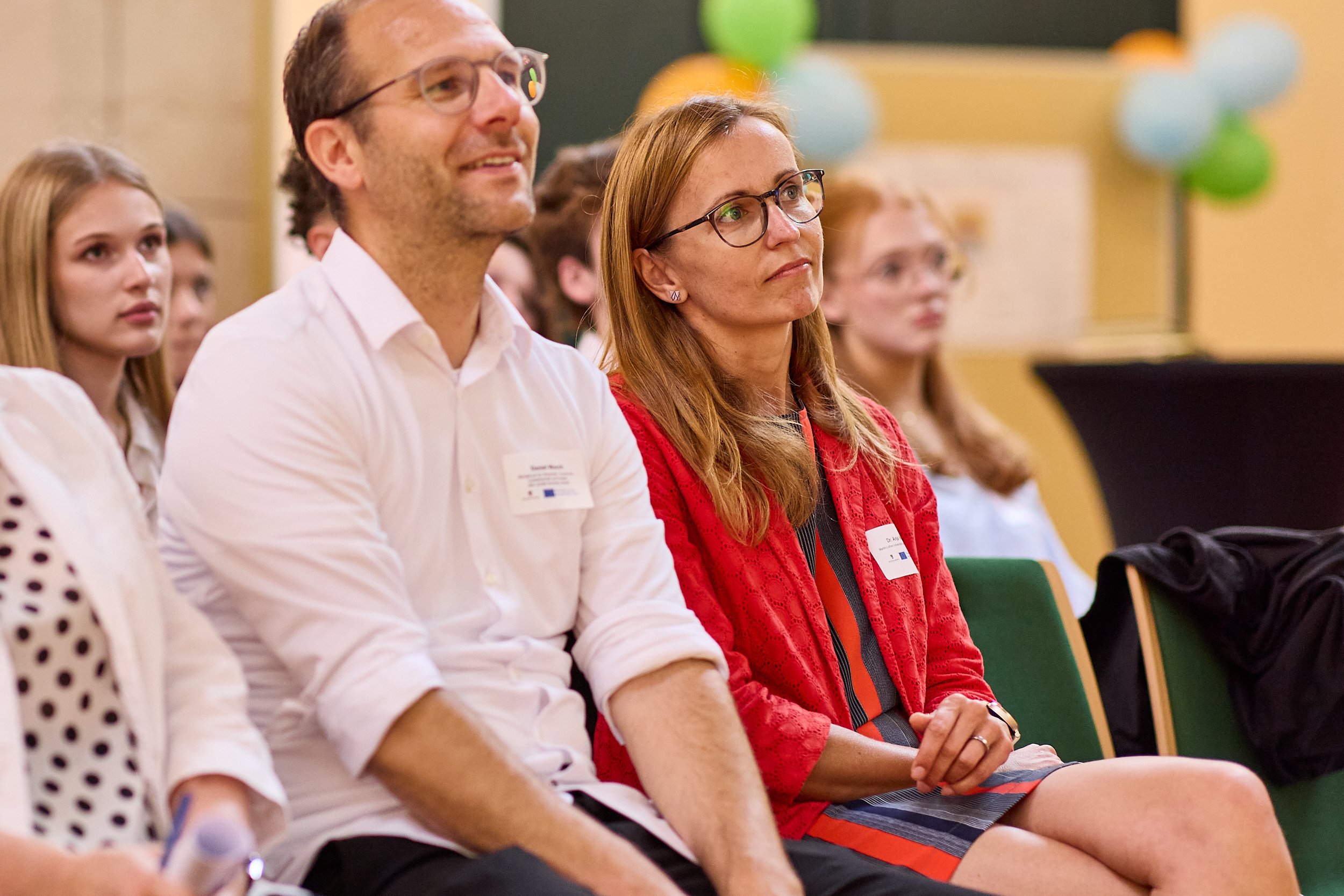 Menschen sitzen in einem Vortragssaal während einer Veranstaltung, die Hauptfokussierten sind ein Mann und eine Frau, beide tragen Namensschilder, die Frau trägt eine rote Jacke und die Männer trägt ein weißes Hemd.