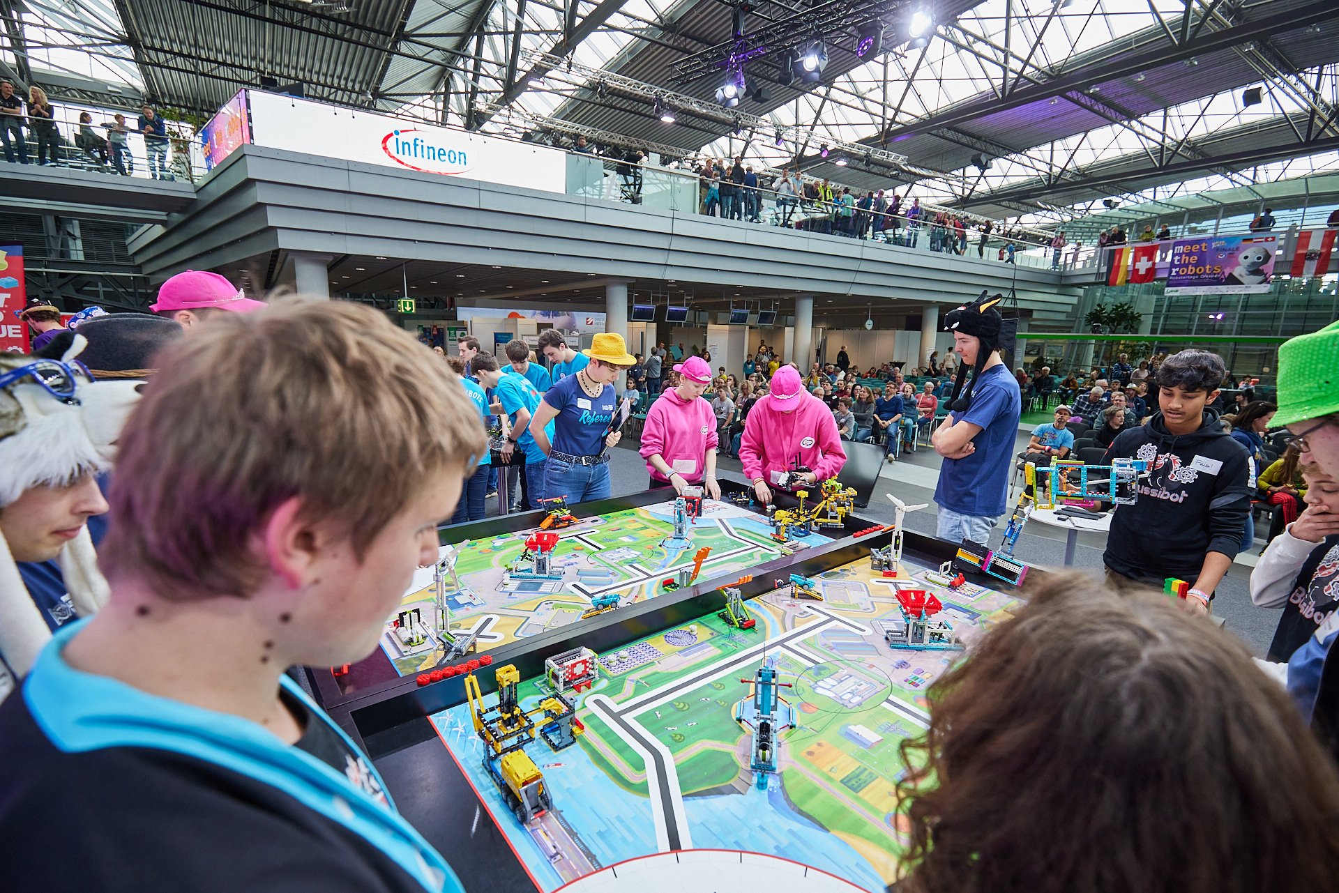 Jugendliche und Kinder spielen mit LEGO-Robotern und bauen an einer Robotik-Schlachtfeld auf einer Messe in einem großen, lichtdurchfluteten Hallenraum mit vielen Zuschauern und mehreren Ebenen.