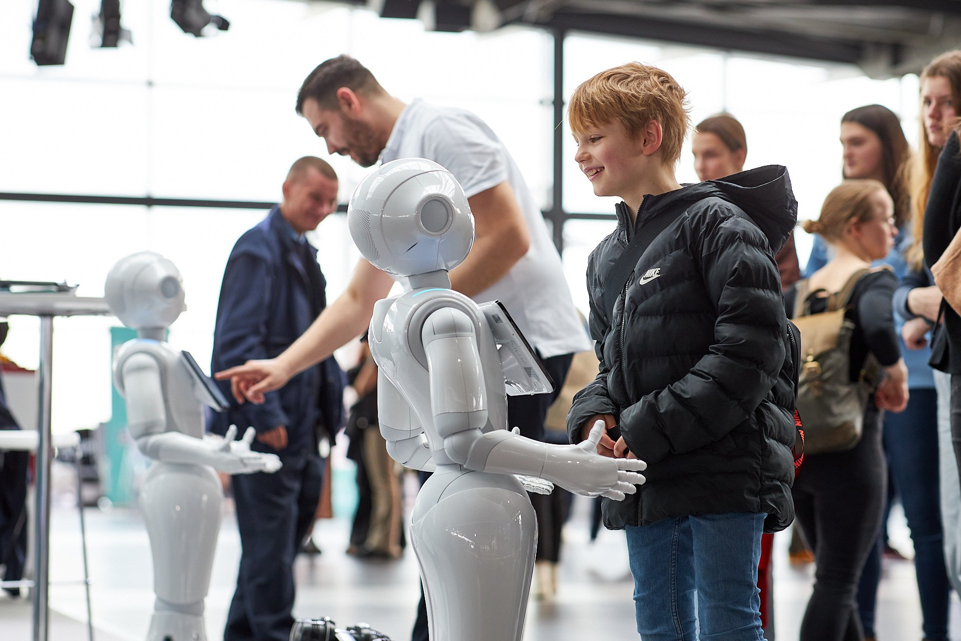 Ein Junge in schwarz-bunter Jacke und blaue Jeans spricht mit einem weißen Serviceroboter mit Tablet im öffentlichen Raum, umgeben von weiteren Menschen und Robotern.