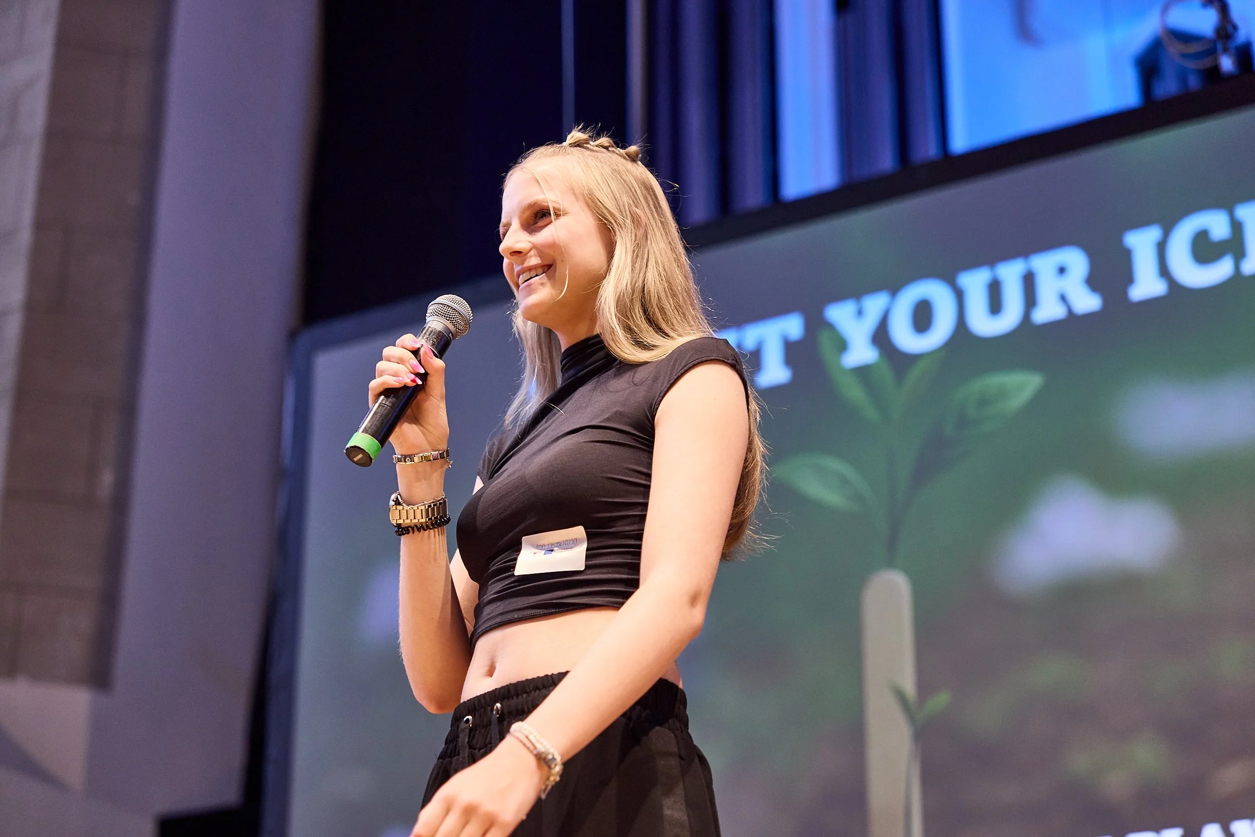 Eine lachende junge Frau mit langem, blondem Haar hält ein Mikrofon und spricht auf einer Bühne. Im Hintergrund ist ein Bildschirm mit dem Text "LET YOUR I" und einem Bild eines jungen Pflänzchens zu sehen.