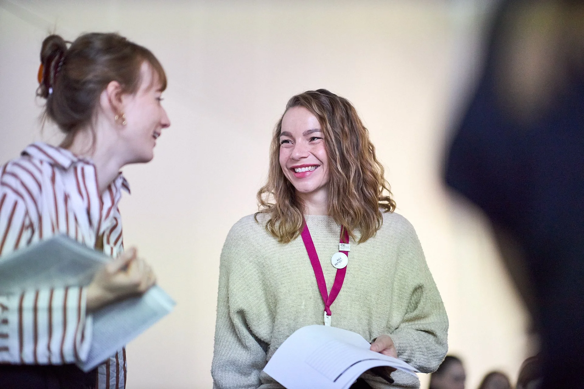 Zwei lachende Frauen, die miteinander sprechen, in einem hellen Raum, eine hält ein Buch, die andere ein Dokument.