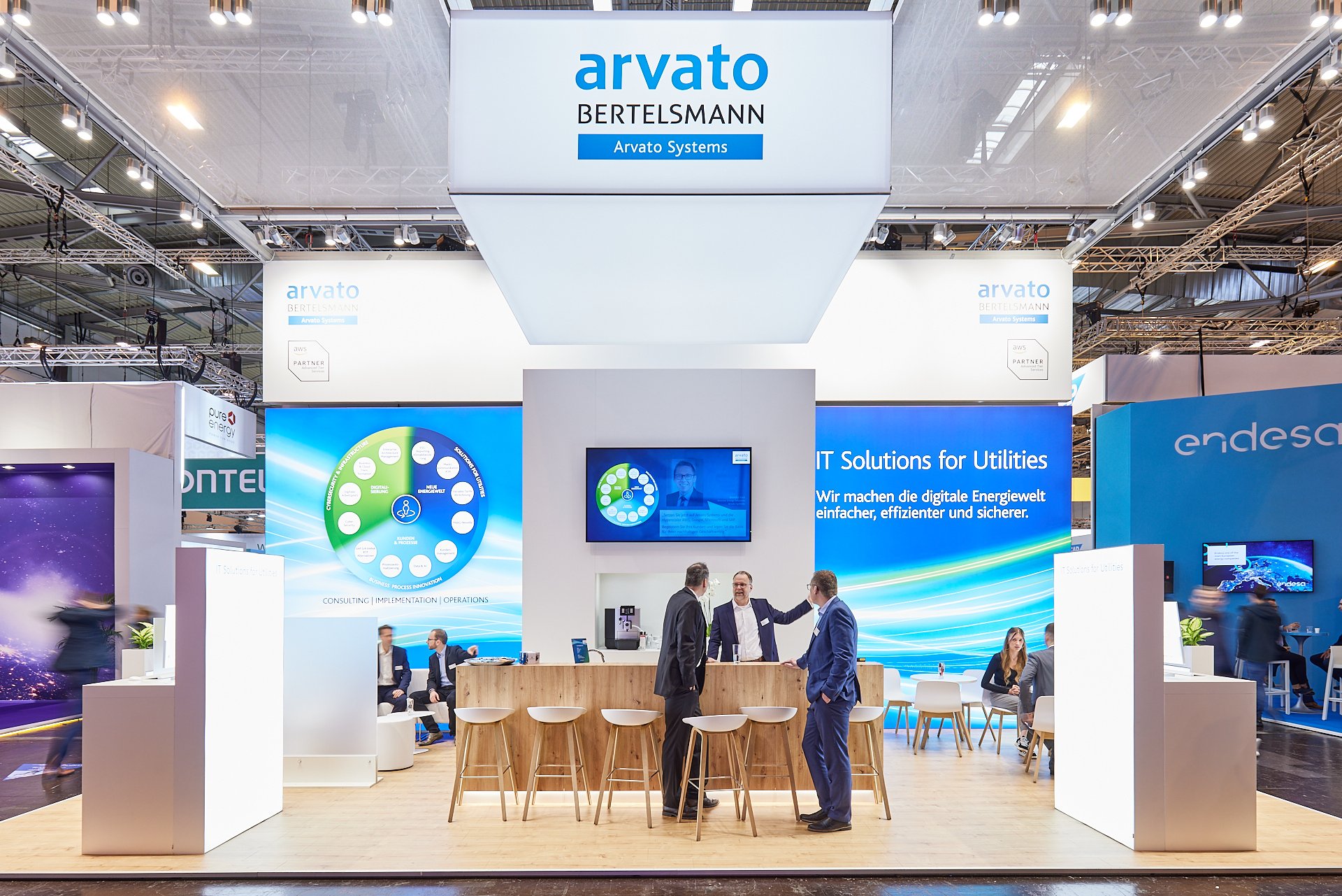Ein Messeausstellungsstand mit mehreren Personen im Gespräch, großen Bildschirmen und Logos für Arvato Bertelsmann und Endesa.