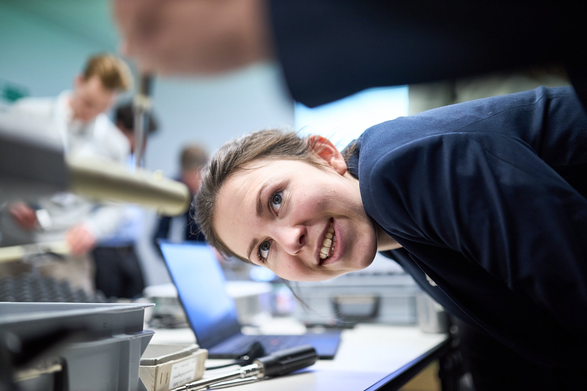 Junge Frau blickt lachend in die Kamera in einem Labor mit anderen Wissenschaftlern im Hintergrund.