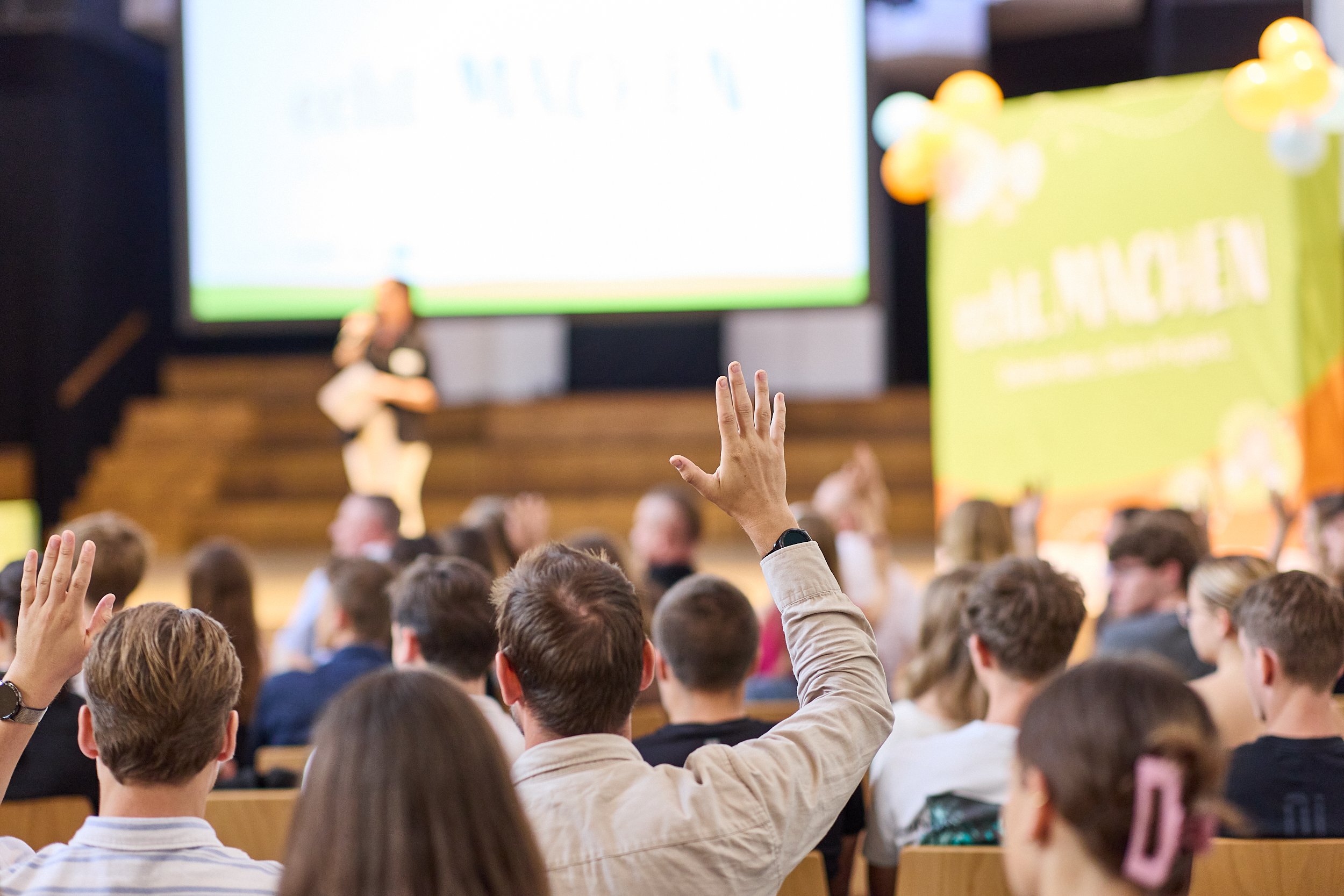 Studenten nehmen an einem interaktiven Vortrag teil, einige heben die Hand, während ein Referent auf der Bühne spricht, im Hintergrund ein Bildschirm und ein grün-gelbes Plakat.