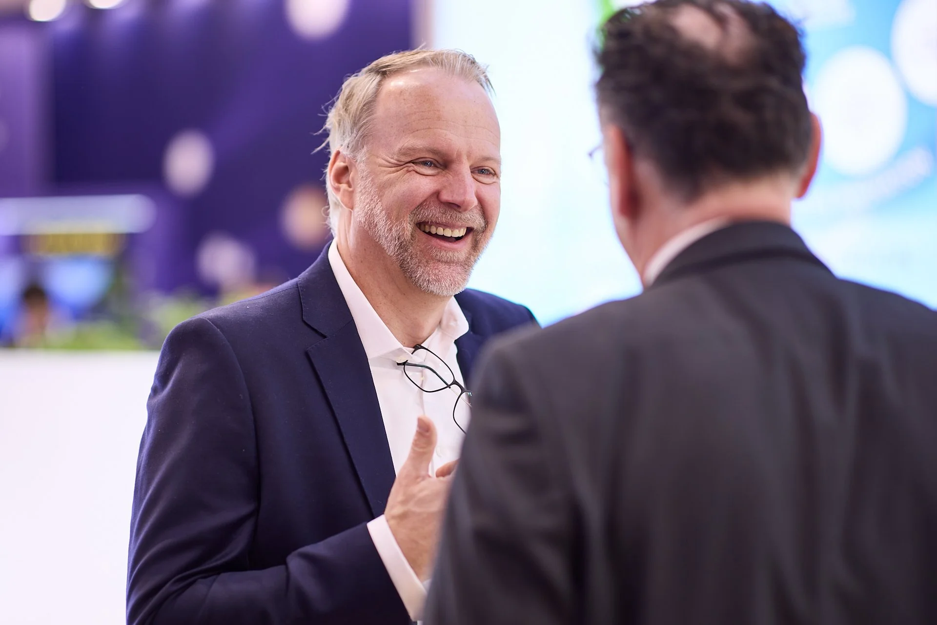 Zwei Männer im Gespräch, einer lächelnd. Der Mann auf der linken trägt einen dunklen Anzug und weiße Hemd, die Brille hängt an seinem Hals. Der Mann auf der rechten trägt einen dunklen Anzug, seine Rückseite ist sichtbar.