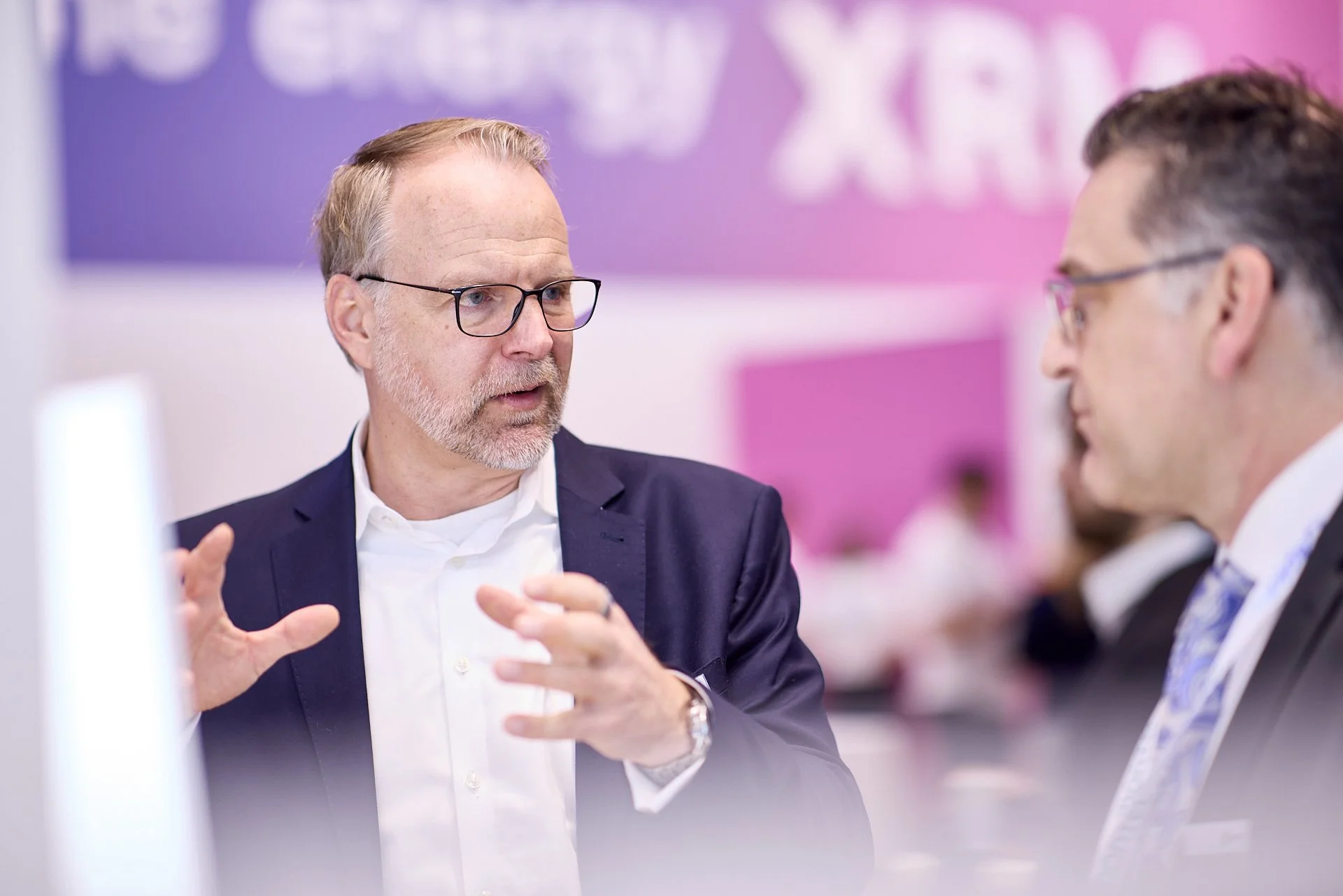 Zwei Männer in einem Gespräch auf einer Konferenz, einer erklärt mit Handgesten, beide tragen Anzüge und Brillen, im Hintergrund pinkfarbige Wand mit Veranstaltungsschild.
