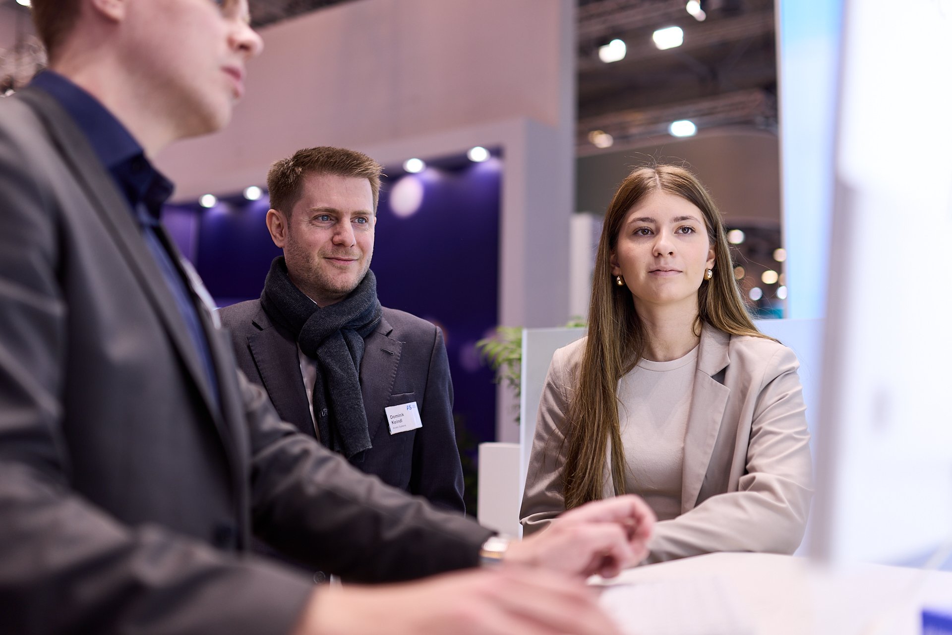 Drei Menschen in formeller Kleidung bei einem Gespräch auf einer Messe oder Konferenz, im Hintergrund mit moderner Beleuchtung und Pflanzen.