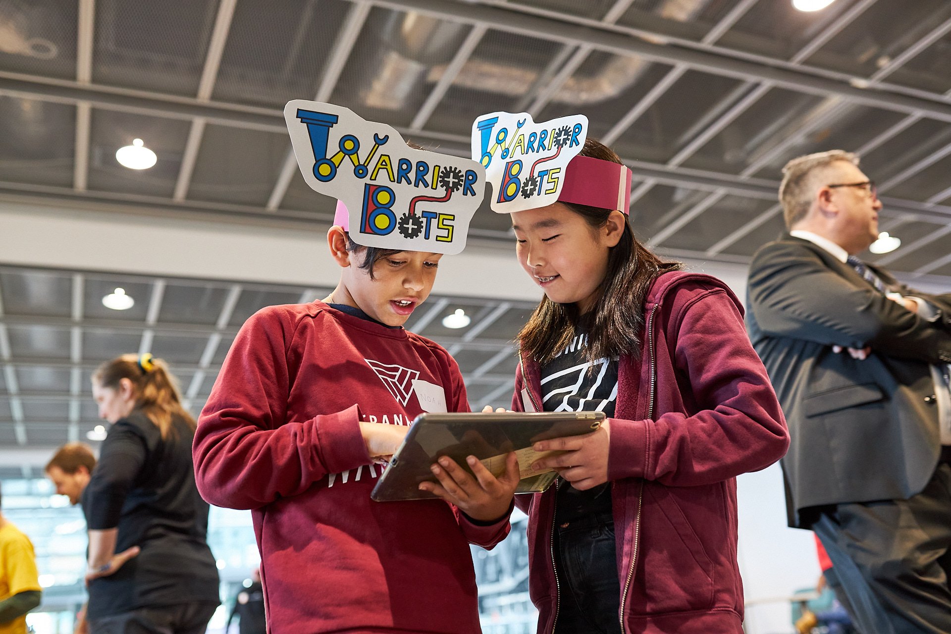 Zwei Kinder tragen bunte Kopfbänder mit der Aufschrift "ToaArrarBOTs" und schauen gemeinsam auf ein Tablet in einer großen Halle, während im Hintergrund andere Personen stehen.