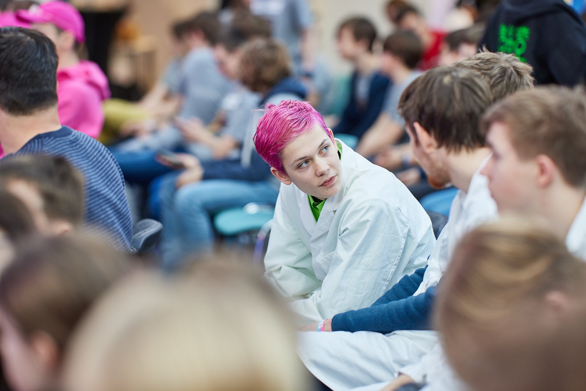 Junge mit pinkfarbenen Haaren sitzt in einer Gruppe von jungen Menschen und spricht mit ihnen in einem Raum.