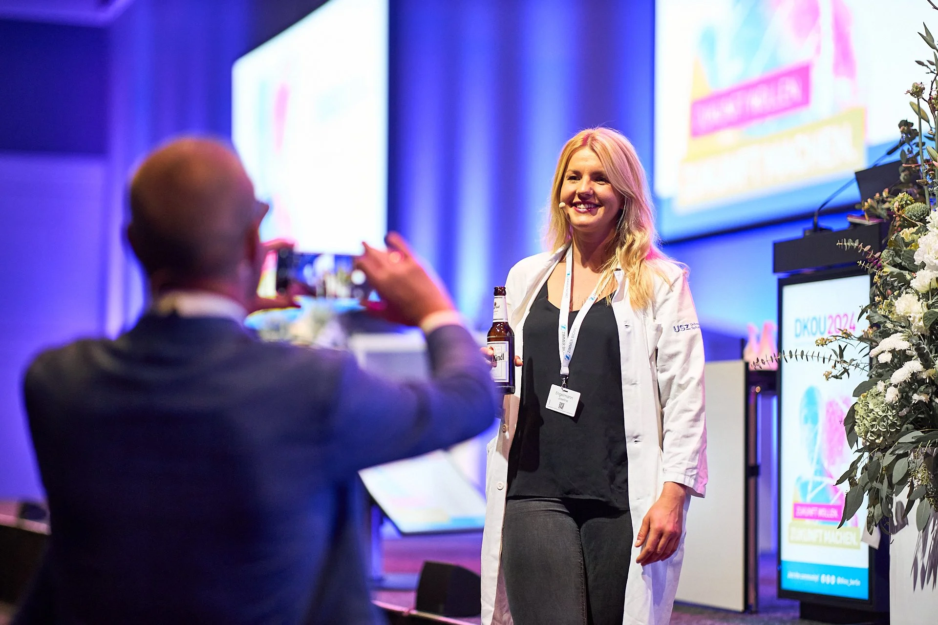 Eine lächelnde blonde Frau in einem weißen Laborkittel posiert für ein Foto auf einer Konferenz. Im Hintergrund sind Bildschirme mit bunten Präsentationen zu sehen.