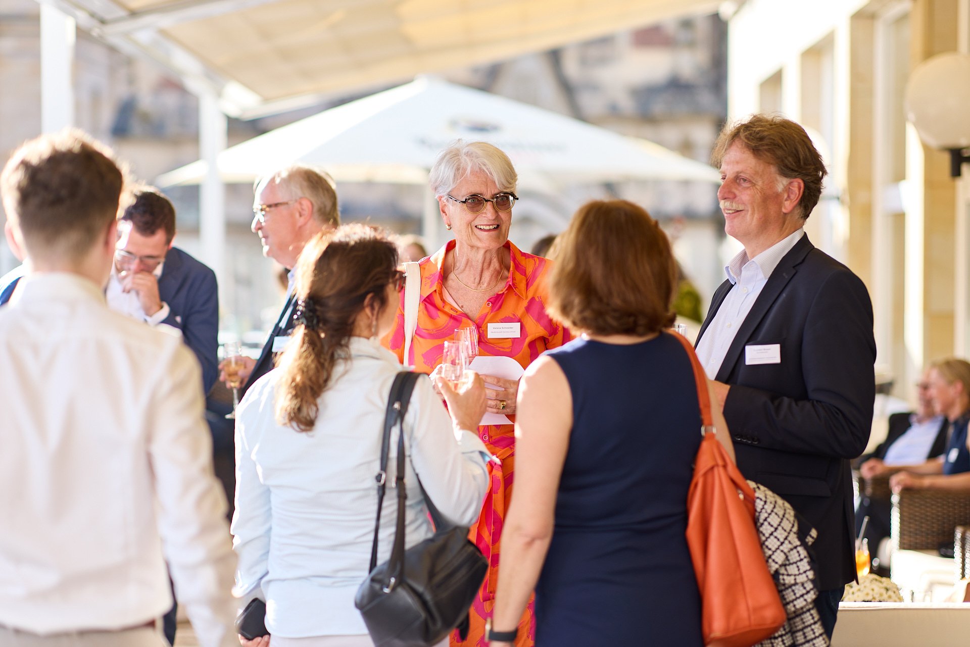 Menschen beim Networking bei einer Veranstaltung, einige halten Gläser mit Getränken, im Hintergrund ein Insektenschutzschirm. 