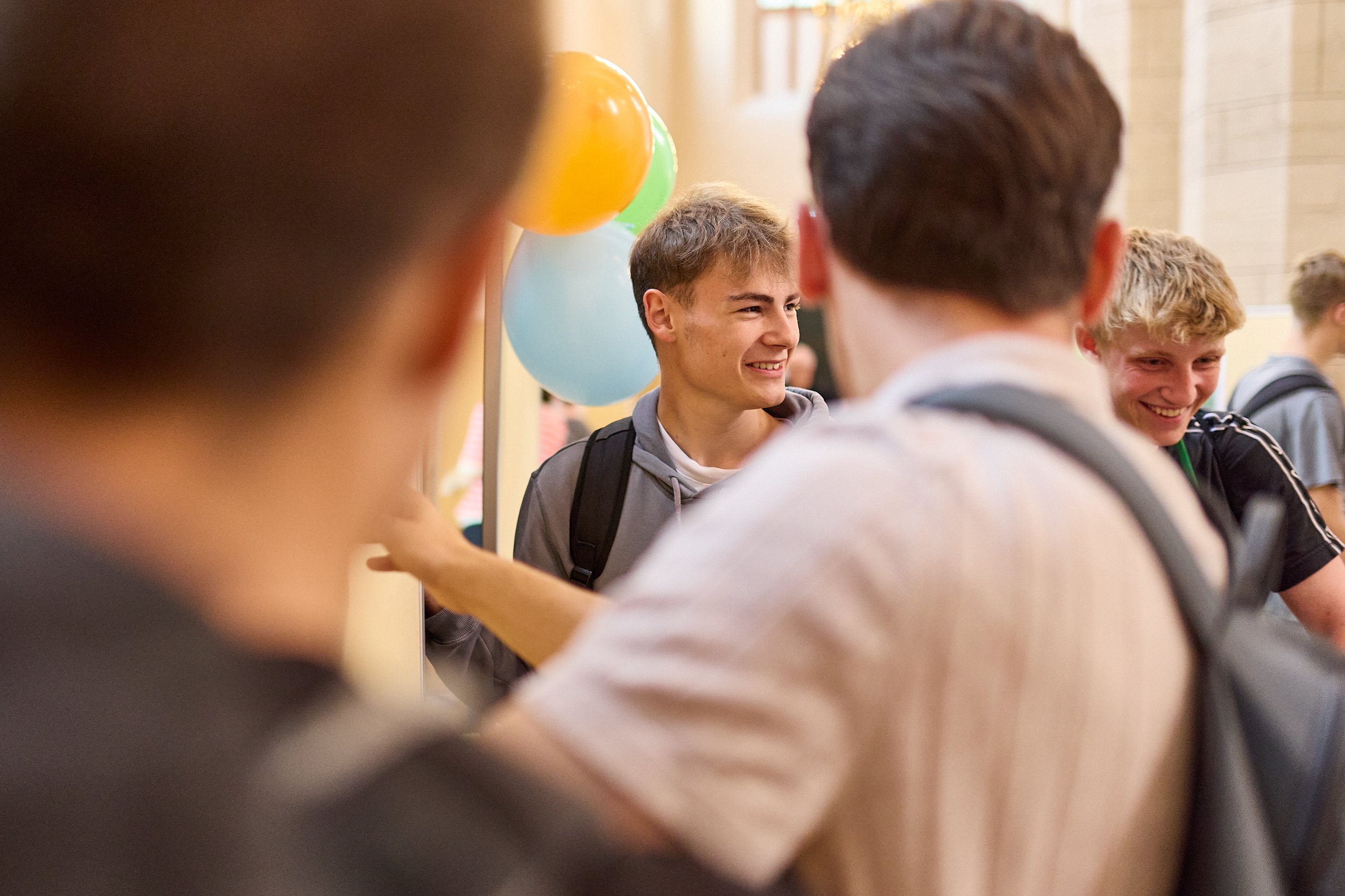 Junge Männer in einem Gespräch, umgeben von bunten Ballons, in einem hellen Raum, vermutlich auf einer Feier oder einem Schulveranstaltung.