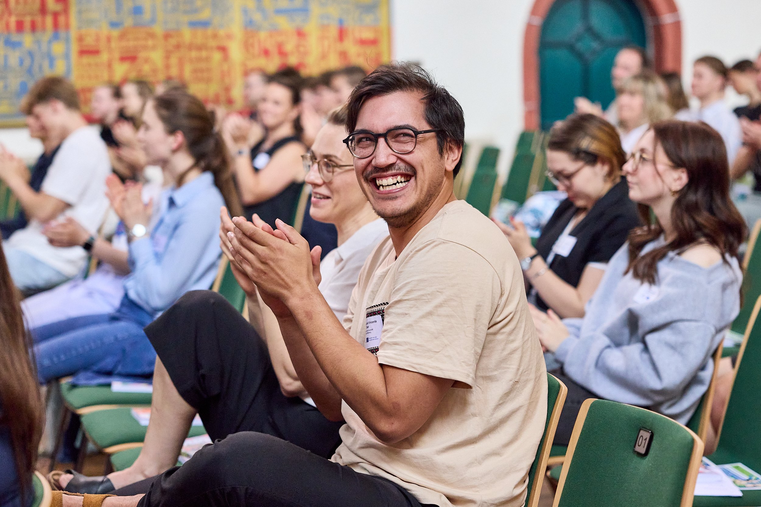 Menschen sitzen in einem Saal, klatschen und lachen, bei einer Veranstaltung oder Konferenz.