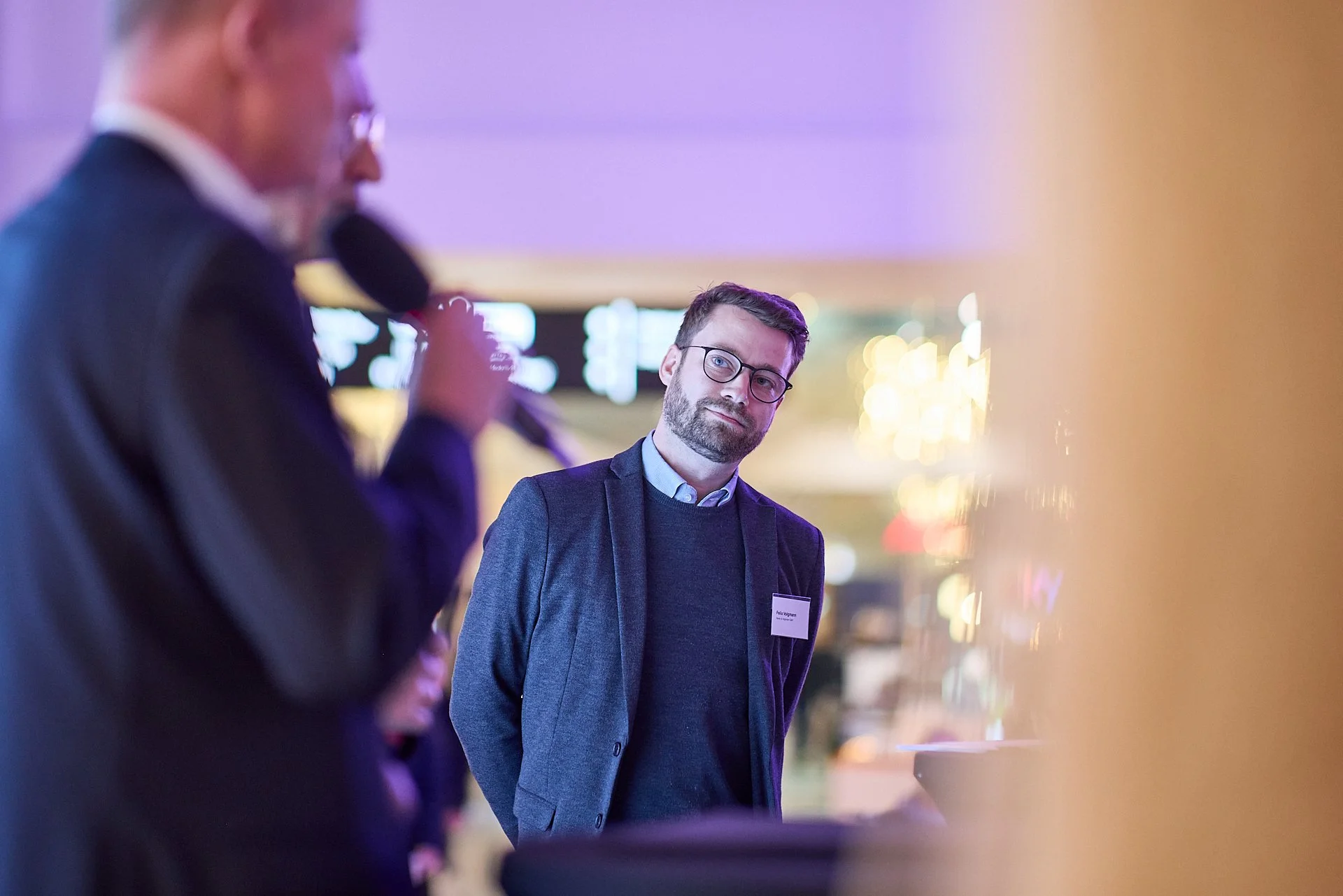 Ein Mann mit Brille in einem dunklen Anzug und hellblauem Hemd hört einem anderen Mann zu, der in einem Anzug an einem Mikrofon spricht, bei einer Veranstaltung in einem gut beleuchteten Raum.