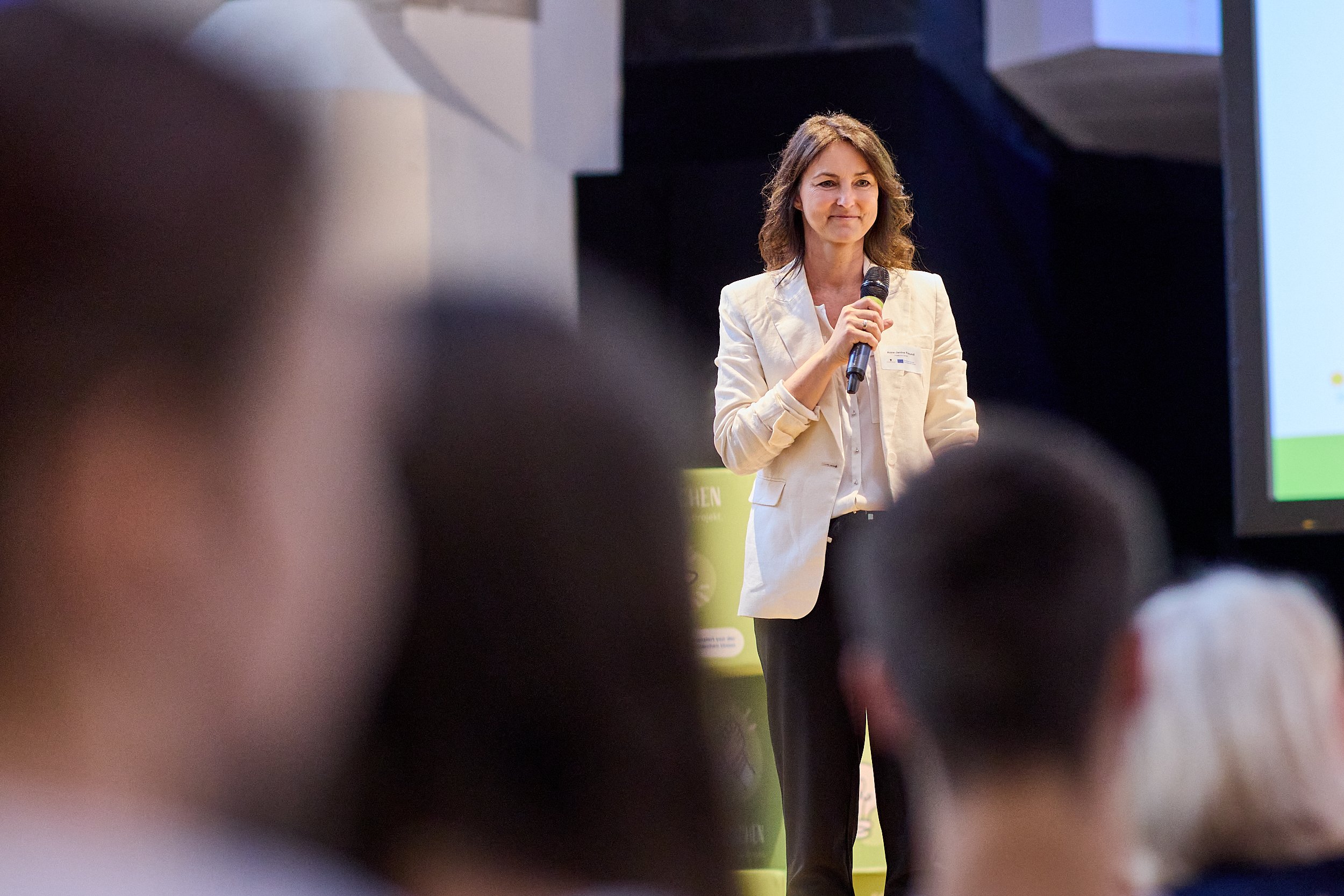 Eine Frau hält ein Mikrofon beim Vortrag vor einem Publikum in einem dunklen Raum.