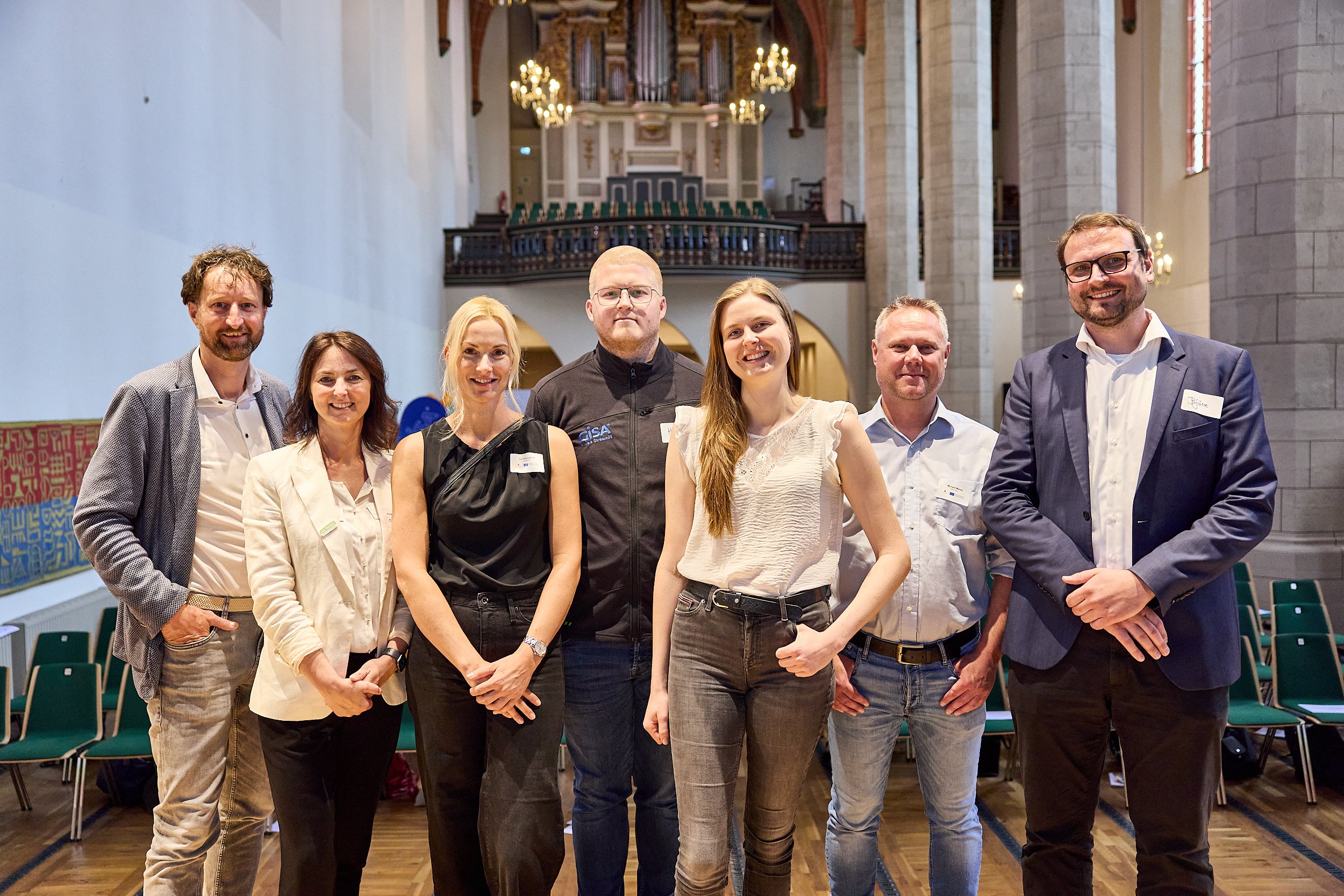 Gruppenfoto von acht Personen in einem historischen Saal mit hohen Fenstern, Holzdecke und Kronleuchtern, die lachen und in formeller Kleidung stehen.