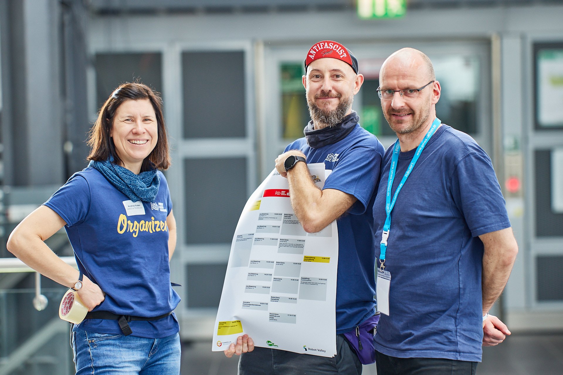 Drei Menschen in blauen T-Shirts stehen nebeneinander und lachen, während der Mann in der Mitte ein großes Programm- oder Zeitplan-Poster hält, im Hintergrund eine moderne Gebäude-Umgebung.