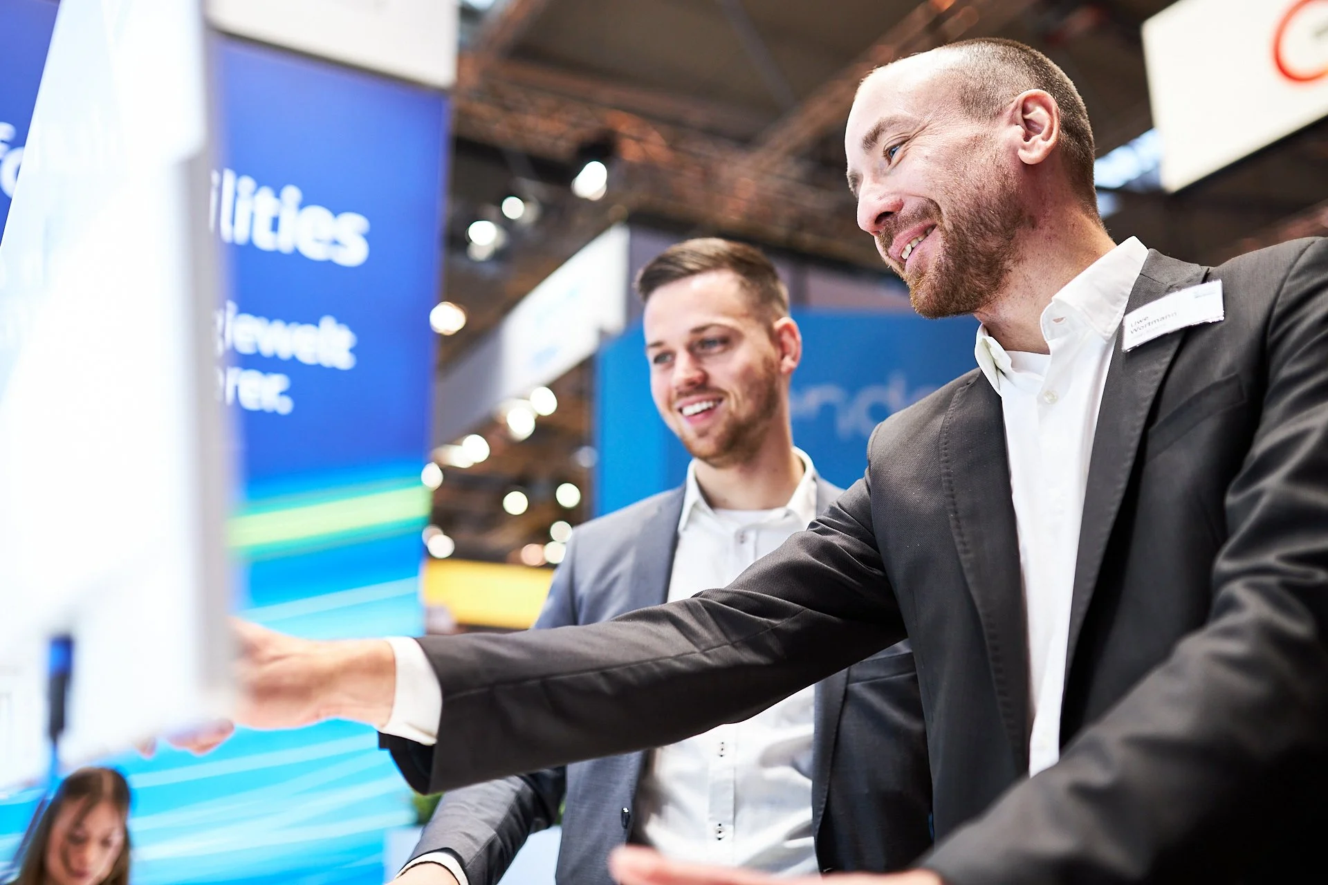 Zwei Männer in Anzügen bei einer Präsentation oder einem Messestand auf einer Konferenz, die gemeinsam auf einen Bildschirm schauen und sich dabei unterhalten.