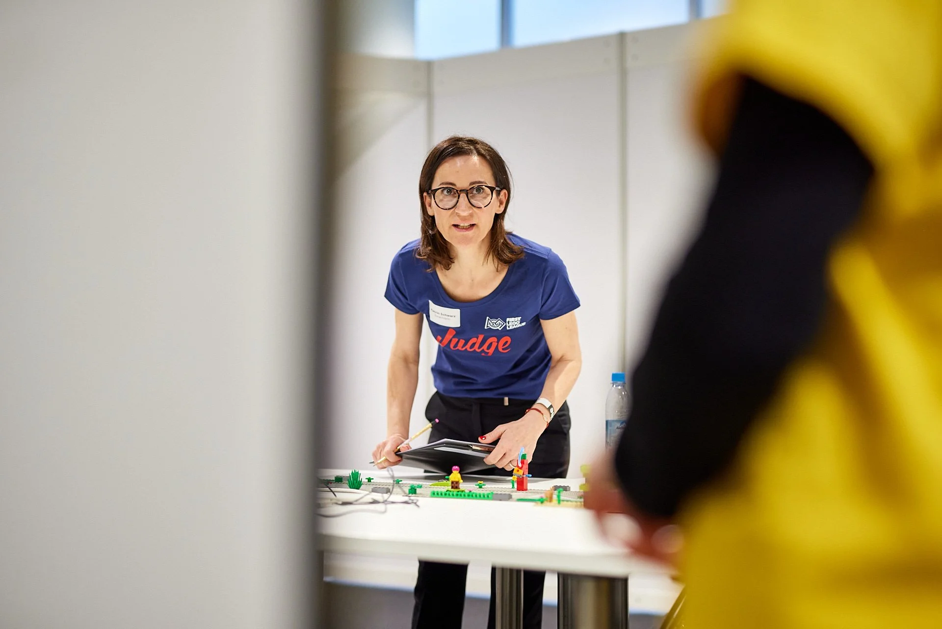 Eine Frau mit Brille und blauer T-Shirt, die an einem Tisch mit Lego-Sets arbeitet, spricht mit einer anderen Person in gelbem Hoodie.