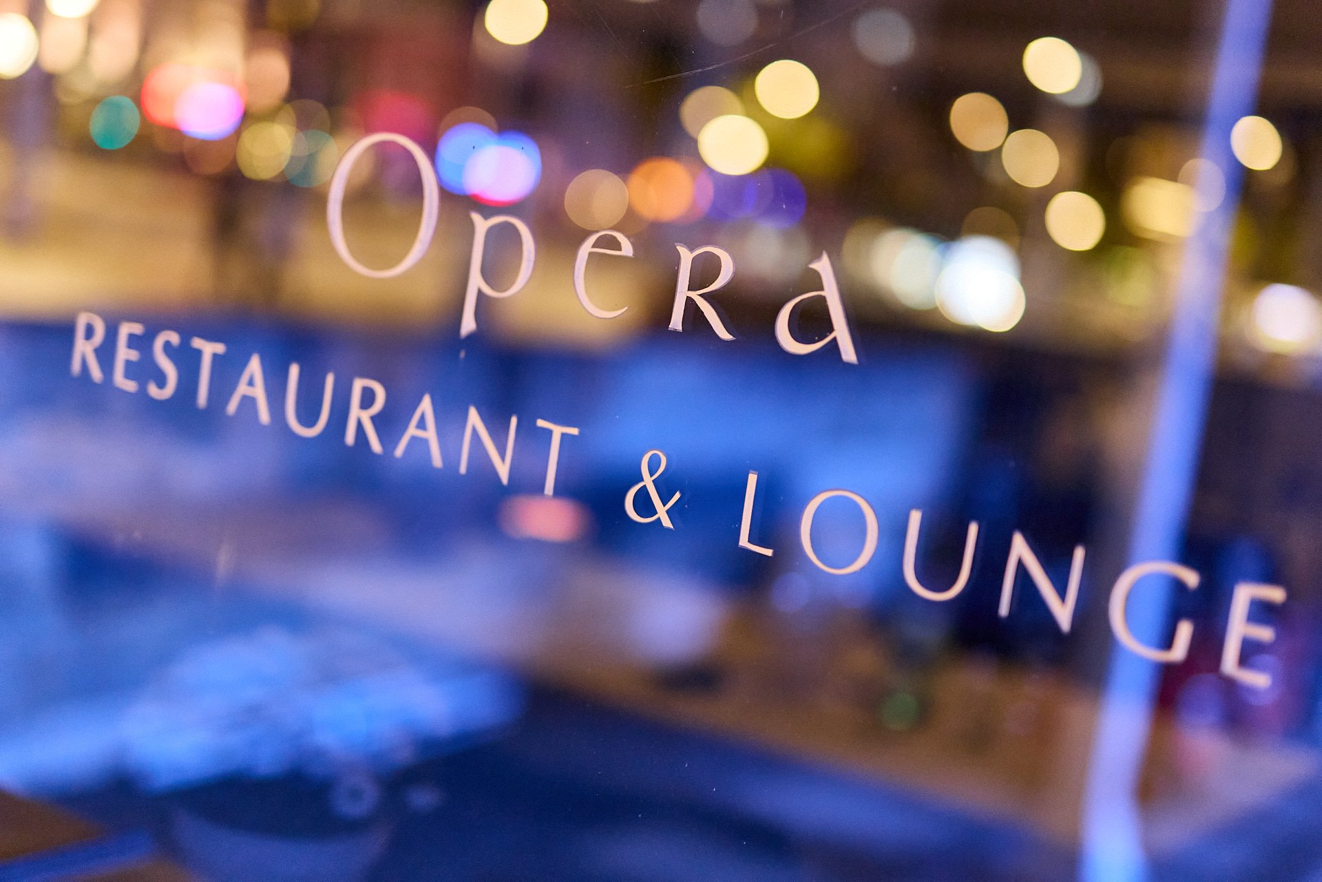 Ein Glas mit der Aufschrift 'Opera Restaurant & Lounge' gegen eine bunte, lichtdurchflutete Stadthintergrund bei Nacht.