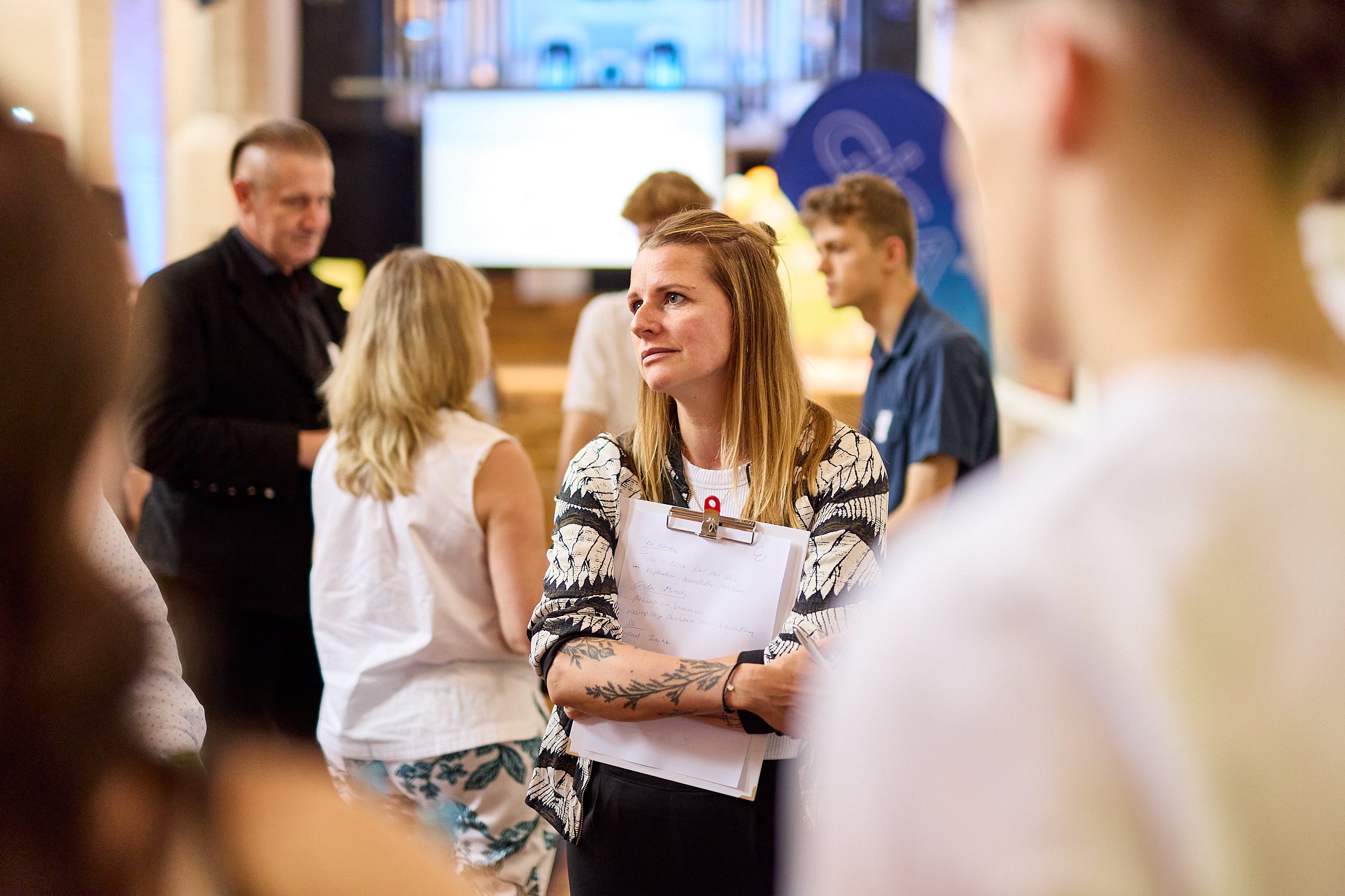 Eine junge Frau mit Tattoos auf den Armen hält einen Aktenordner und zeigt ein nachdenkliches Gesicht. Sie steht in einer Gruppe von Menschen bei einer Veranstaltung.